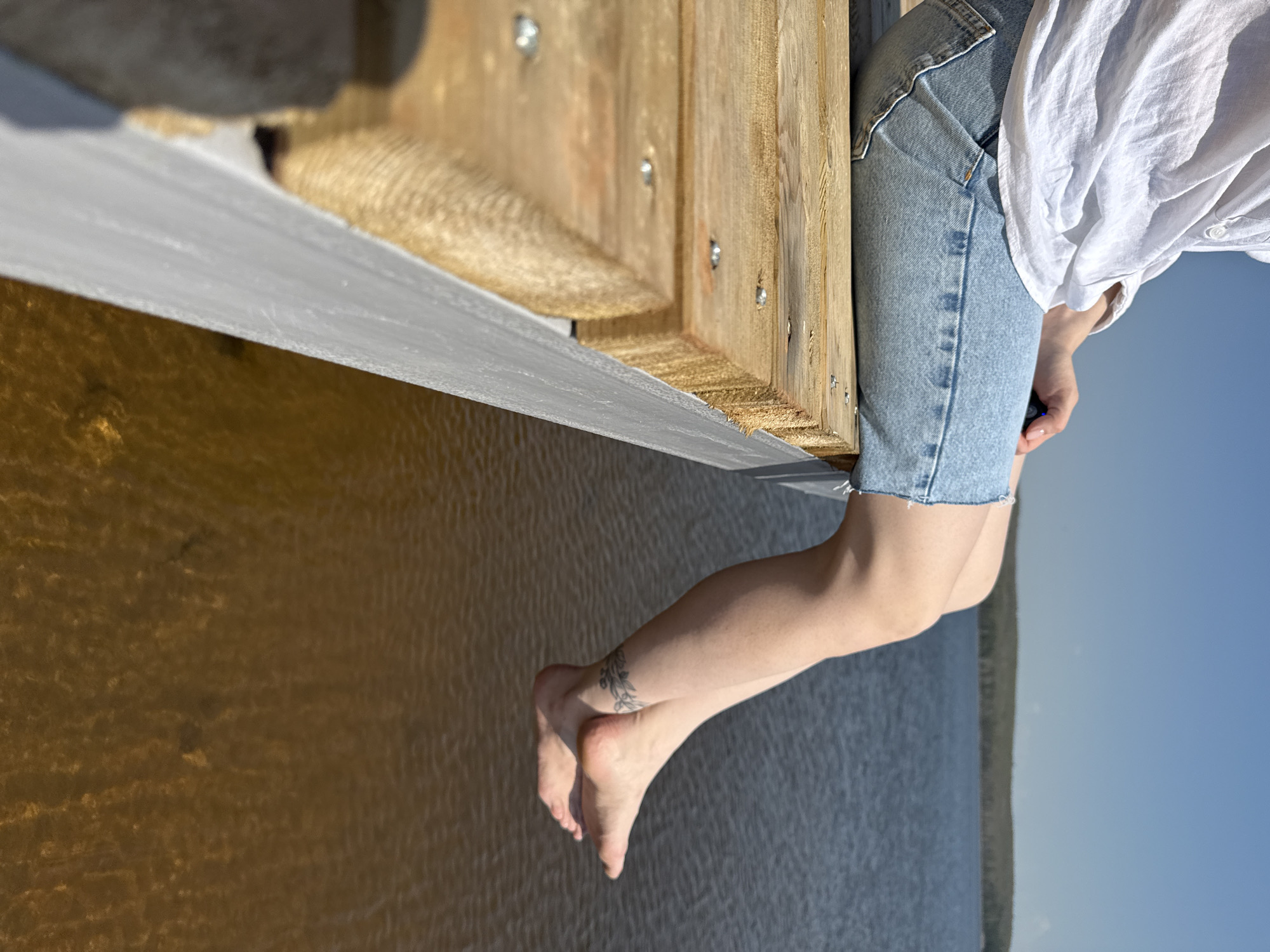 A person wearing denim shorts and a white top is sitting on the edge of a wooden structure overlooking a body of water under a clear blue sky. The location could be near Chaikovsky, Russia, given the context. The person's legs are dangling, and a tattoo is visible on one ankle. The wooden structure appears to be part of a pier or dock, with a metallic trim on its edge. The water is brownish-yellow and possibly shallow.