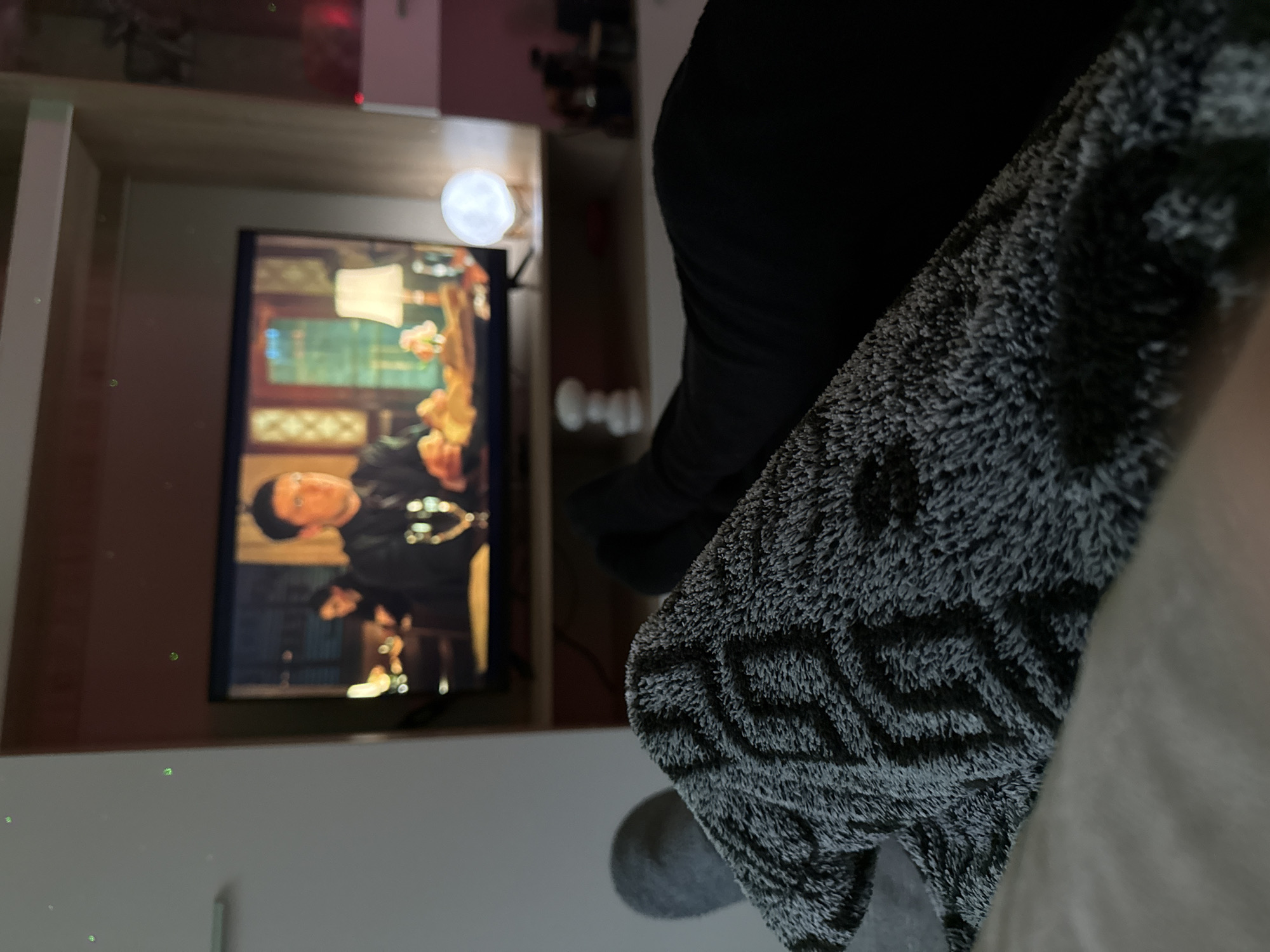 A close-up, rotated indoor shot shows a television playing a program featuring a man with black hair and a black suit, presumably in a residence. The TV is mounted within a light-colored cabinet or shelving unit. Obscuring a portion of the view is a dark-colored, possibly patterned blanket or fabric. In the periphery, out-of-focus objects are visible, and the lighting is soft and diffuse, suggesting an interior scene. The location is indeterminable from the given information.