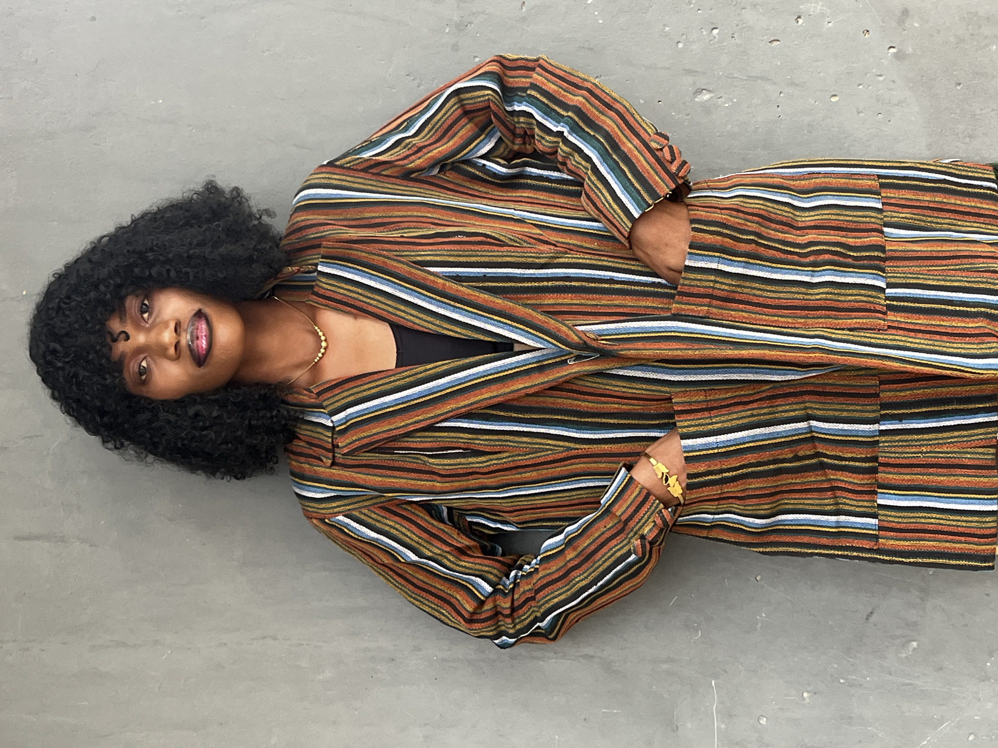 A medium shot shows a woman with dark skin, facing the camera at an angle while lying on a gray concrete surface. Her curly black hair contrasts with her gold necklace and bracelets. She wears a striped blazer and matching pants featuring horizontal stripes of various colors, including orange, brown, blue, and white. A small portion of a black top is visible under the blazer. The setting appears to be an urban outdoor area, possibly in the city of Badagry, Nigeria, based on context. The surface she lies on is cracked and uneven, suggesting an outdoor environment.