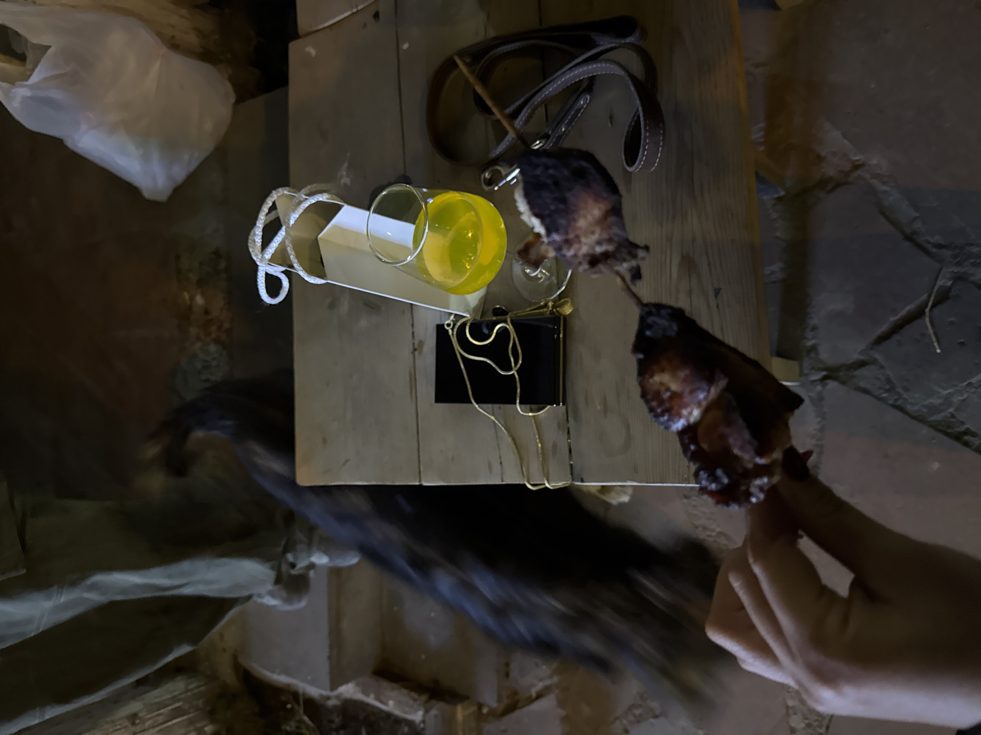 The image shows a table cluttered with various items. There's a glass of yellow liquid, a black purse with a gold chain, a brown leather strap, and a white bag. Skewered meat is being held by a hand in the lower right corner. The table is made of light-colored wood and is set against a backdrop of stone-like material. The location is Yerevan, Armenia, but there is no identifying feature that connects the location with the image.