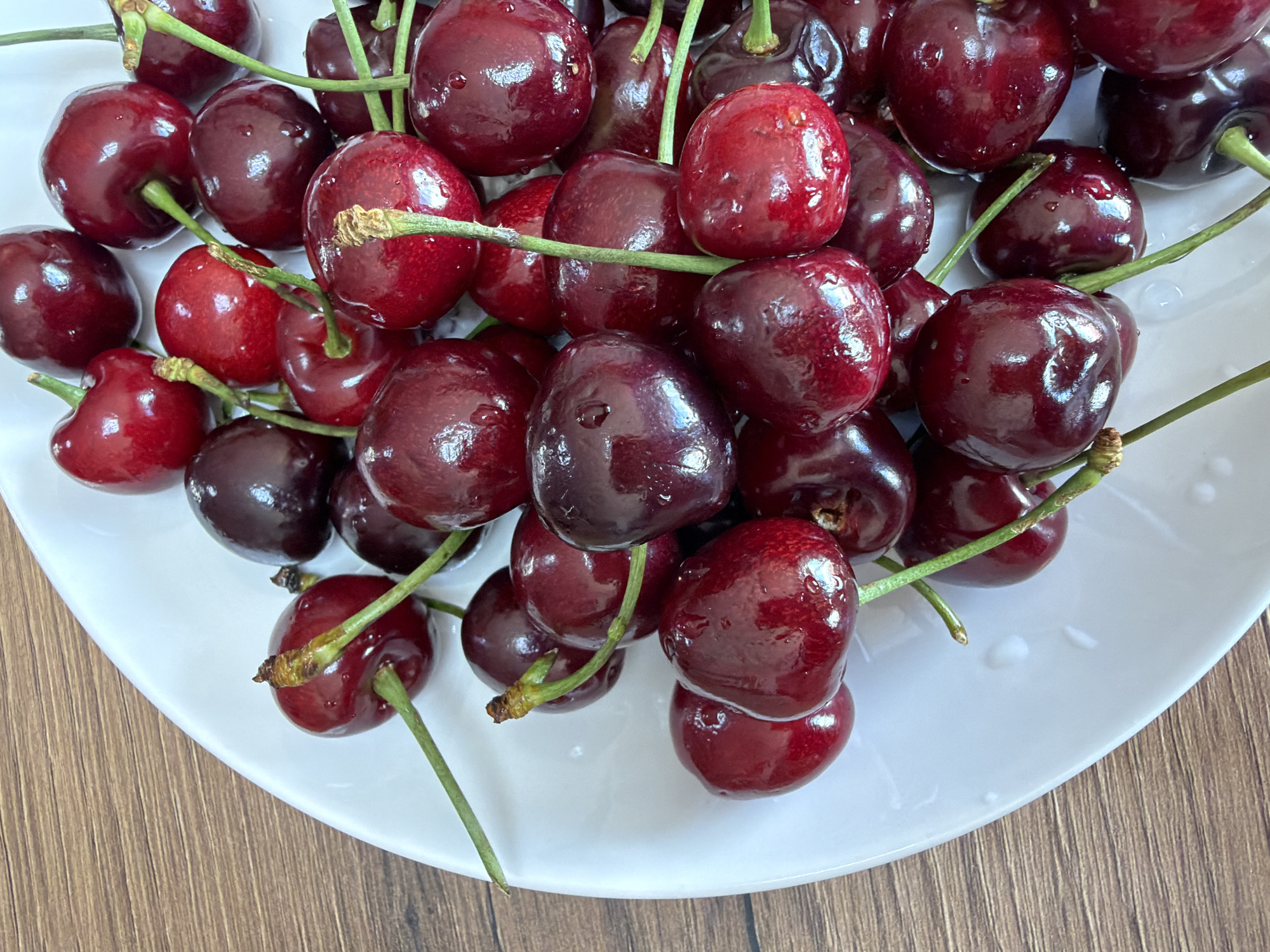 A close-up, overhead shot shows a white plate filled with a pile of ripe, dark red cherries. The cherries are glossy and some have water droplets on them, indicating they may have been recently washed. They are mostly round with some slightly heart-shaped. The stems of the cherries are green and some are still attached. The white plate is round and has a slightly textured surface with subtle indentations. The plate is resting on a wooden surface with a visible grain pattern. The image is well-lit, emphasizing the vibrant color and texture of the cherries. There are no people or animals present in the image, and no visible signs of the city of Chaikovsky, Russia.