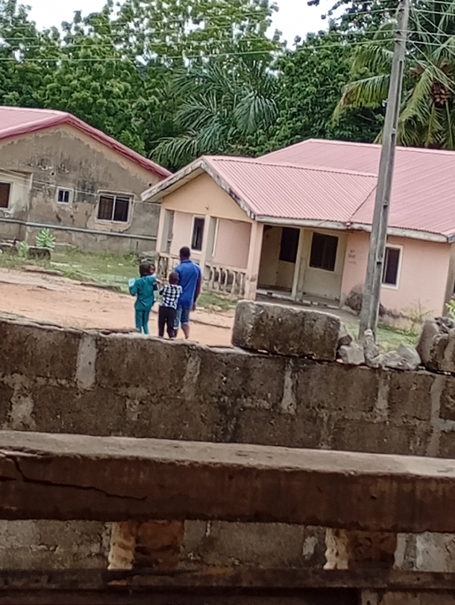 The image shows a partially visible brick wall in the foreground. Behind the wall, three individuals are visible: an adult male wearing a blue shirt and shorts, and two children, one in a blue outfit and the other in a checkered shirt. They appear to be walking near two single-story buildings with red roofs. The building on the left is older and more weathered. Behind the buildings, there is dense green foliage, including what appears to be palm trees. An utility pole is visible to the right of the building with the red roof. The location is presumed to be in a residential area within the city of Unknown, Unknown.