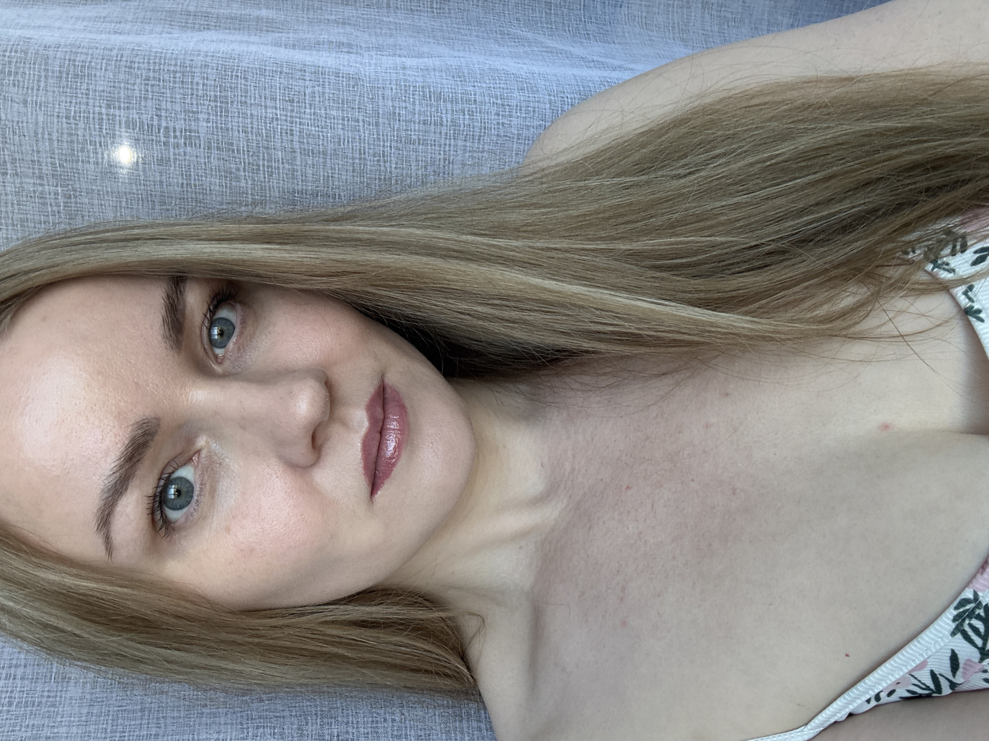 A fair-skinned woman with long blonde hair is pictured. She is wearing lipstick and a floral-patterned garment. The background is a textured, light-colored fabric. The location is not determinable from the image.