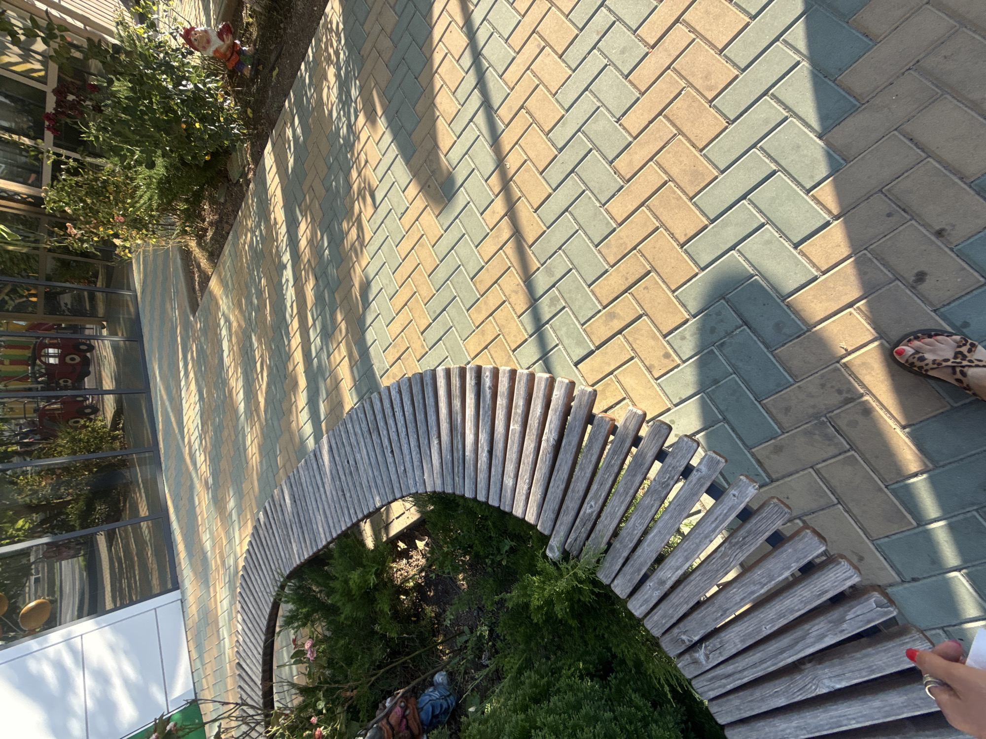 The image depicts an outdoor scene in the city of Зуйское сельское поселение, Ukraine. In the foreground, a person's feet are visible, wearing leopard-print sandals, standing on a paved walkway. The walkway is laid with rectangular bricks in a herringbone pattern, alternating between tan and light green colors, and is partially covered by dappled sunlight and shadows. To the left, a curved wooden bench with a weathered finish encloses a small garden bed filled with green bushes and a few pink flowers. A garden gnome with a red hat and holding something in its hands is partially visible near the bushes. In the background, a modern building with large reflective windows is situated, showing reflections of trees and what appears to be playground equipment. The overall scene suggests a public outdoor area, possibly a park or recreational space, in the specified Ukrainian location.