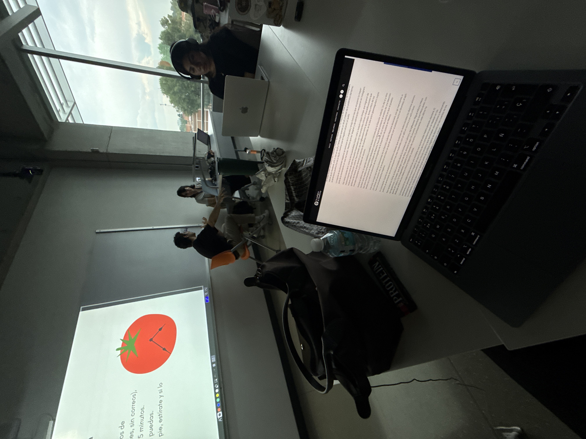 A medium shot shows several people working in a brightly lit room in Mexico City, Mexico.
People: Several individuals sit at desks working on laptops. One person near the window wears headphones. Another individual stands at the whiteboard.
Subjects: The primary focus is on a laptop in the foreground, displaying text on its screen. A whiteboard shows a graphic of a tomato-shaped clock with hands pointing to a time. A graphic shows a tomato clock is labeled "os de", "es, sin correos)," "5 minutos.", "puedas.", "pie, estírate y si lo"
Objects: Laptops, desks, chairs, a water bottle, a bag, and a book titled "PROTEIN" are visible. The scene takes place near a window.
Actions: People are working on their laptops. One person appears to be teaching or presenting something on the whiteboard.
Interactions: The interaction is mostly individual, with each person focused on their work.
Environment: The room is brightly lit, possibly a study hall or classroom. The background shows an urban landscape through the window, hinting at the location in Mexico City. The weather appears cloudy.
