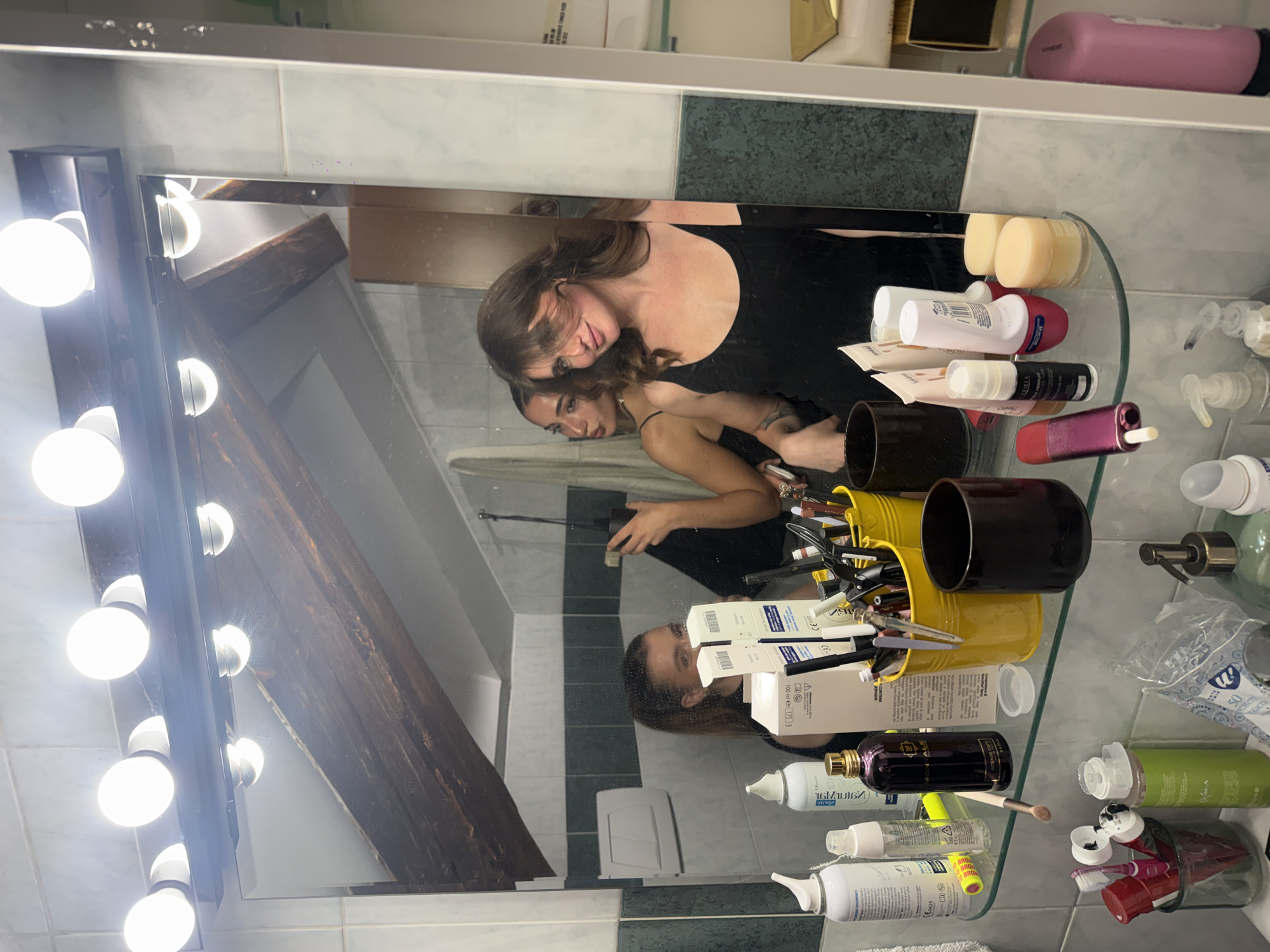 Here is a detailed description of the image:
Scene: The image appears to be a reflection in a bathroom mirror.
People: A woman is visible in the mirror, seemingly taking a photo of herself or checking her appearance. She is wearing a black top. Her reflection is also visible, creating a double image effect.
Objects: The mirror has multiple lights attached to its frame. Various cosmetic and personal care products are arranged on a shelf in front of the mirror. These include bottles, jars, makeup brushes in containers, tubes of creams or lotions, and a package of wet wipes.
Environment: The bathroom has tiled walls with a combination of light and dark green tiles. There is a wooden beam or structure visible in the reflection, suggesting a particular architectural feature or decor element.
Details:
- The lighting is bright, likely due to the vanity lights.
- The arrangement of products on the shelf seems somewhat cluttered.
- The woman's focus seems to be on her reflection, possibly applying makeup or grooming.
Location: While it's challenging to definitively confirm the location in the city of Milan, Italy, based solely on the image, the architectural details such as the tiled walls and visible wooden beam might be indicative of certain styles or building types found in older residential areas of Milan. However, without additional contextual information, pinpointing the exact location remains speculative.
