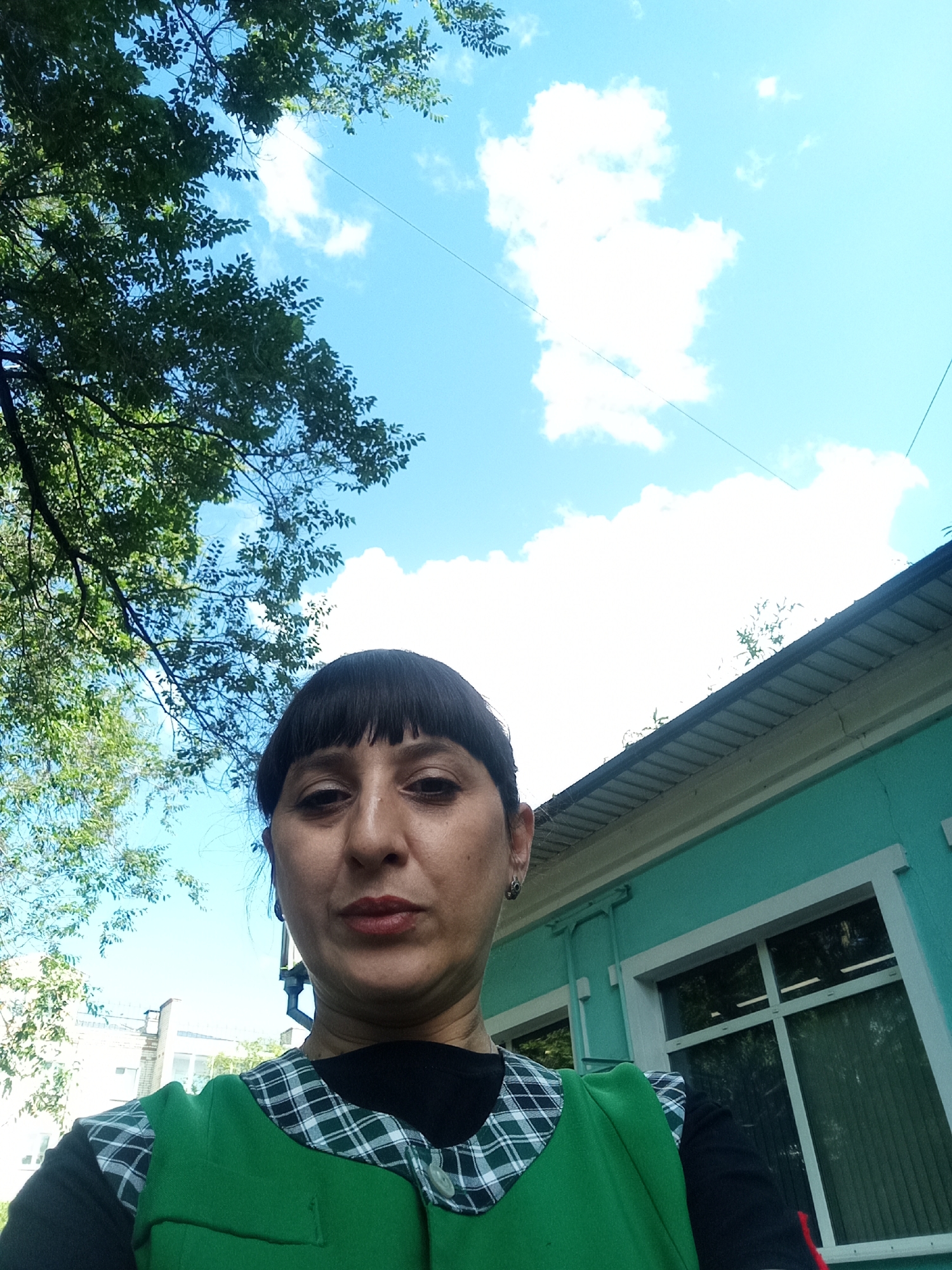 A woman with dark hair and bangs is looking at the camera in a selfie. She is wearing a green vest over a black shirt with a white and black checkered collar. Behind her, the sky is blue with white clouds and some green trees. To the right of the woman is a teal building with a silver roof and white trim around the windows. The image appears to have been taken outdoors during the daytime.