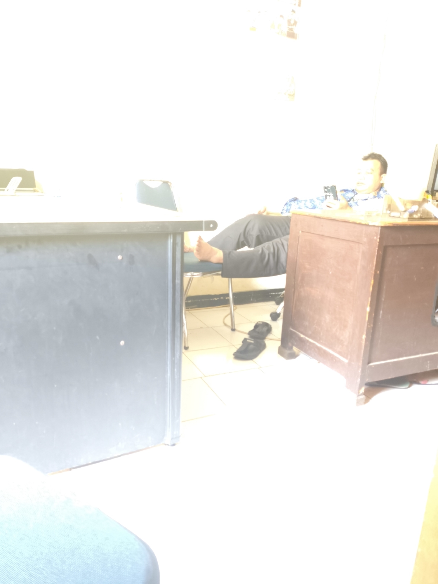 A man is sitting at a desk in what appears to be an office. He is wearing a blue and white patterned shirt and black pants. His legs are propped up on a chair. He is looking at his phone. There are two desks in the image, one dark gray on the left and a brown wooden one on the right. There is a chair with wheels next to the brown desk. A pair of black sandals are on the floor next to the chair. The lighting in the image is very bright, almost overexposed. The scene takes place indoors, likely in an office in Cilegon, Indonesia.