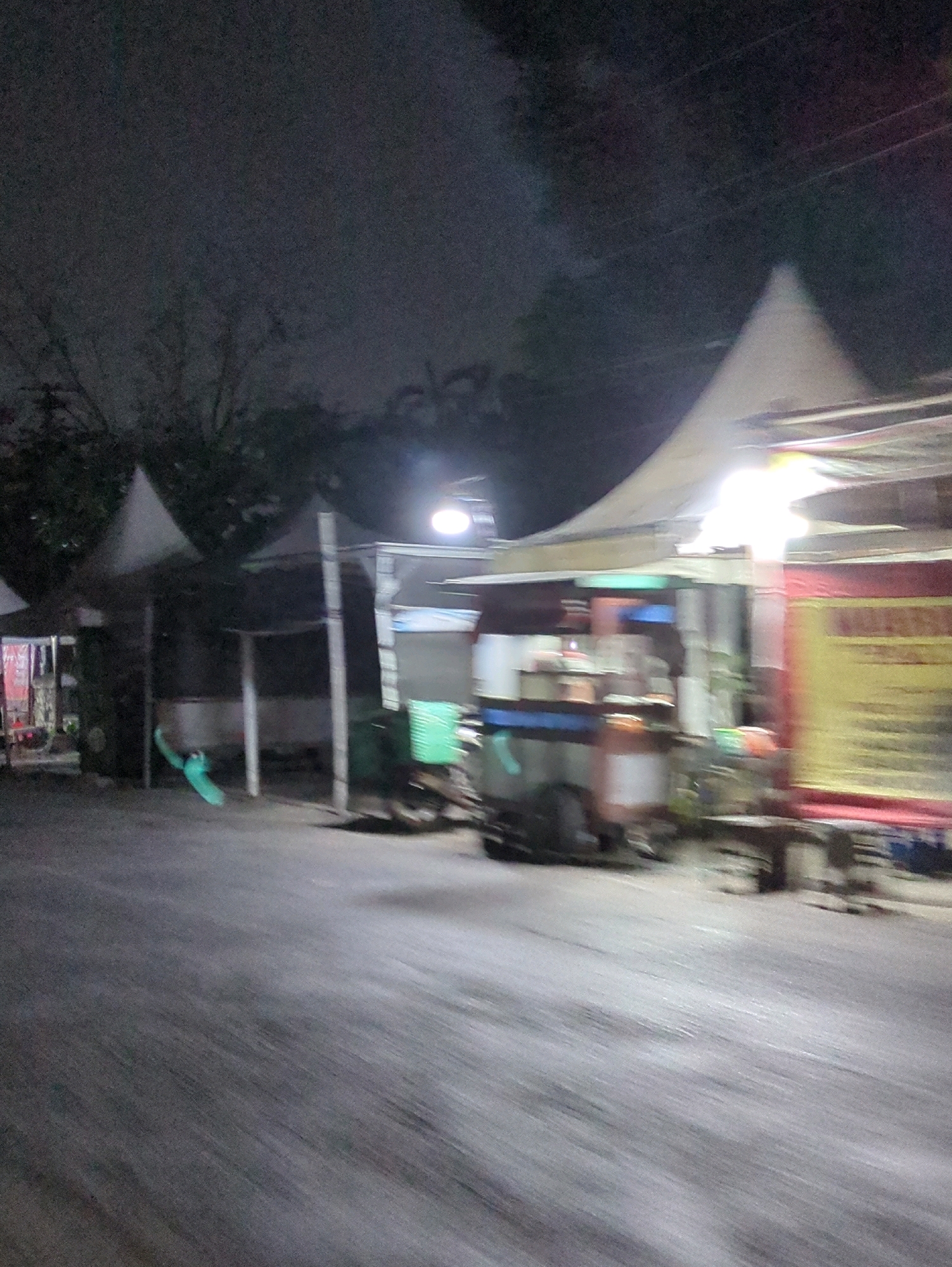 The image depicts a nighttime scene of an outdoor market or food stalls in Colomadu, Indonesia. The foreground shows a blurred asphalt road, suggesting movement. Behind the road, several market stalls with tent-like awnings are visible. One stall on the right is brightly lit and has a yellow sign with red lettering, partially obscured by motion blur. Another stall to the left appears to have a food cart or vehicle with various items displayed. Trees and a dark sky with some cloud cover form the background. The overall atmosphere is that of a bustling, open-air market after dark.