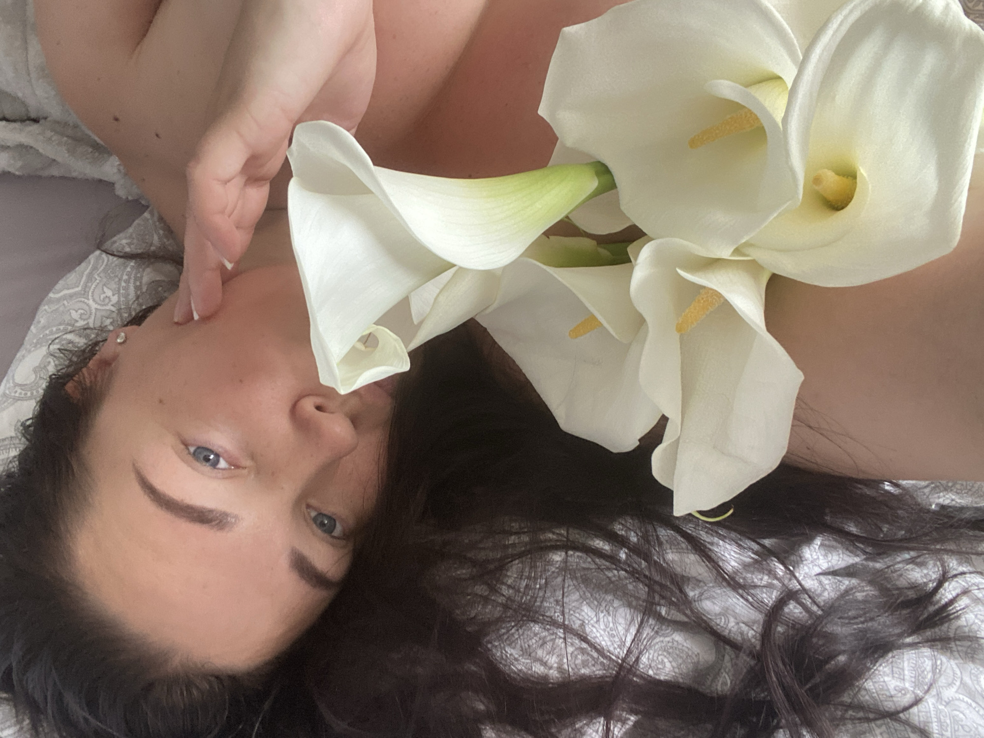 The image shows a woman lying in bed, holding several white calla lilies. She has long, dark hair and is wearing light-colored clothing. The bed is covered with patterned linens. The woman's face is partially visible, and she is looking towards the camera. The image appears to be taken indoors, likely in a bedroom. There is no indication of the location being the city of Sochi, Russia.