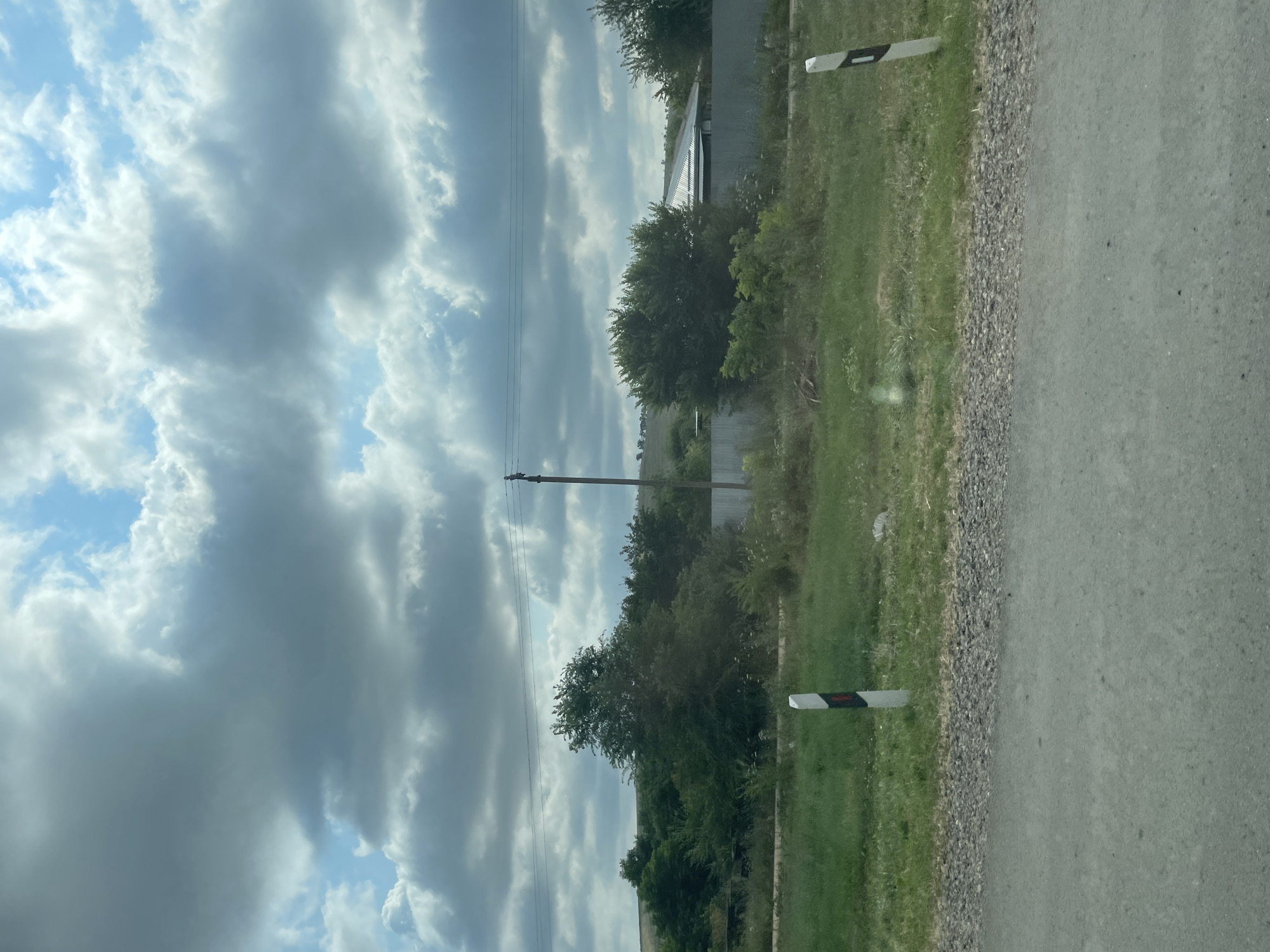 The image shows a rural landscape in the city of Donskoye, Russia. The foreground is a gray asphalt road, bordered by a strip of gravel. Beyond the gravel, a grassy area is visible, with two white posts with black and red markings along the side of the road. In the middle ground, a metal fence runs horizontally, behind which are trees, some bushes, and a building with a metallic roof. A single utility pole with wires stretches across the frame. The background consists of rolling hills covered in vegetation and a sky filled with large, gray and white clouds.