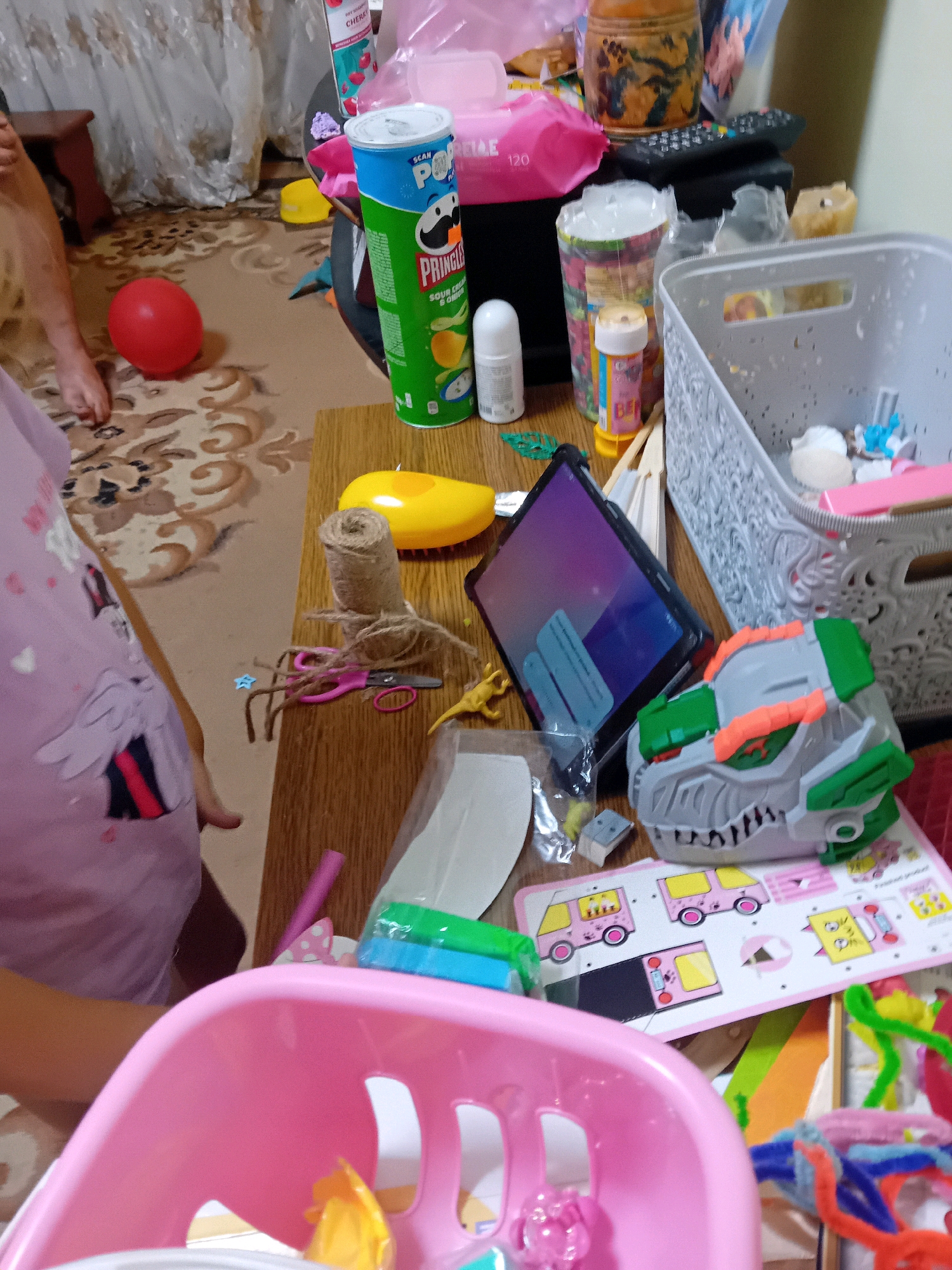 The image depicts a cluttered tabletop, likely in a residential setting in Chuhuiv, Ukraine. The tabletop is laden with various items, including a Pringles can, a roll of twine, scissors, a yellow duck-shaped toy, a tablet, a toy dinosaur gun, and a grey plastic basket filled with small objects. In the foreground, a pink plastic toy basket is visible. A red balloon lies on the floor to the left of the table. A child's arm and torso, wearing a pink t-shirt with a unicorn design, are partially visible on the left side of the frame. The background shows a curtain and a floor with a patterned rug. The overall impression is one of a messy, child-occupied space.