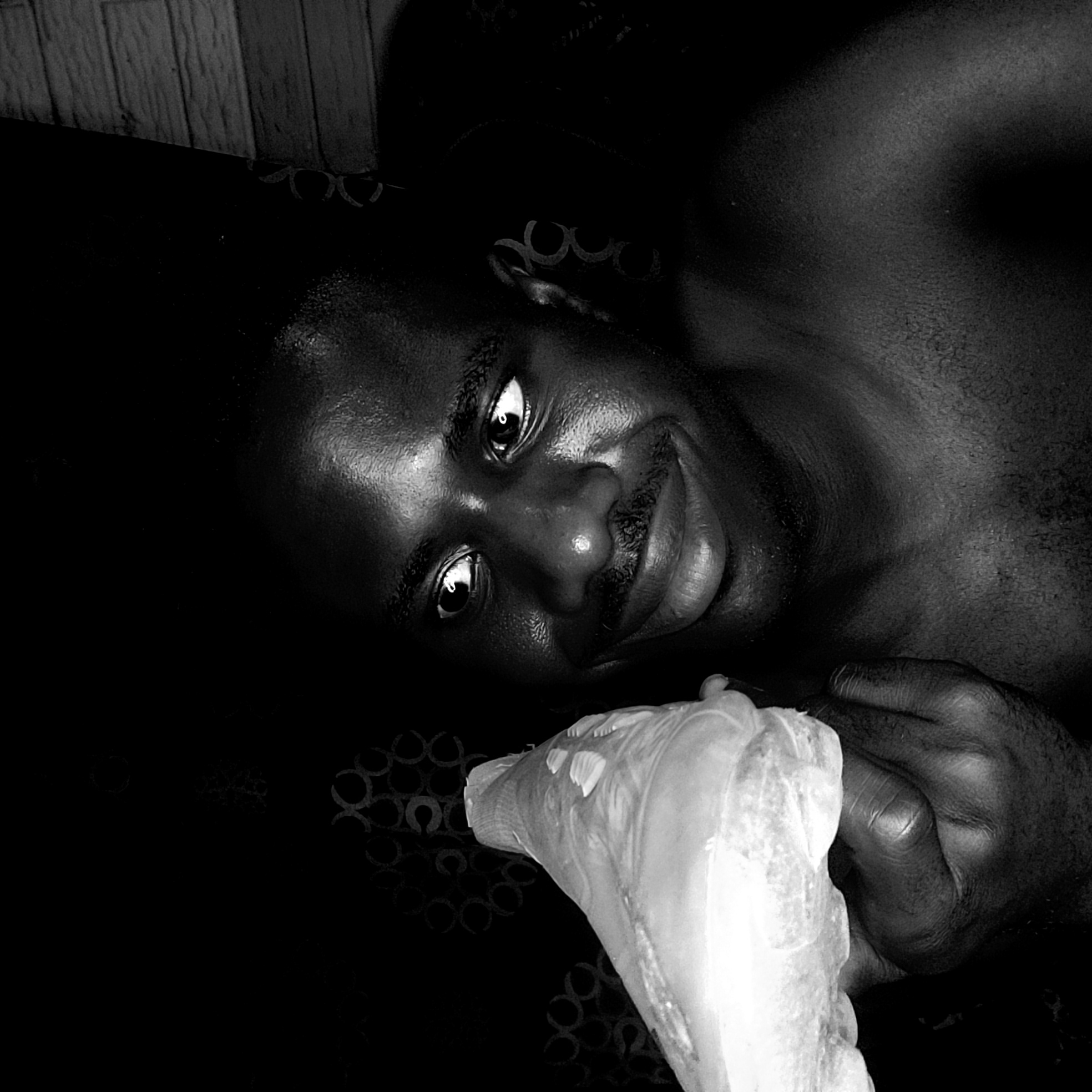 A black and white, close-up portrait shows a person, likely male, lying down and looking directly at the viewer with a slight smile. The individual has dark skin, wide eyes with visible reflections, and a mustache. Their left hand is prominent in the foreground, holding a large, light-colored, irregularly shaped object that resembles a piece of food, possibly a chicken wing or a similar item.
The scene is set indoors, likely in a bedroom or a cozy living space, suggested by the relaxed posture and background elements. In the upper left, a diagonal section of wooden planks or paneling is visible. The dark background features subtle, repetitive circular patterns on what appears to be fabric, possibly bedding or upholstery. The lighting is low and dramatic, creating strong contrasts and highlighting the texture of the skin. The person's expression appears calm and engaged, possibly conveying curiosity or a gentle amusement. There is no visible text. The time of day is not discernible, but the low light conditions suggest an evening or night setting. The image offers an intimate view of the subject and their interaction with the object they hold.