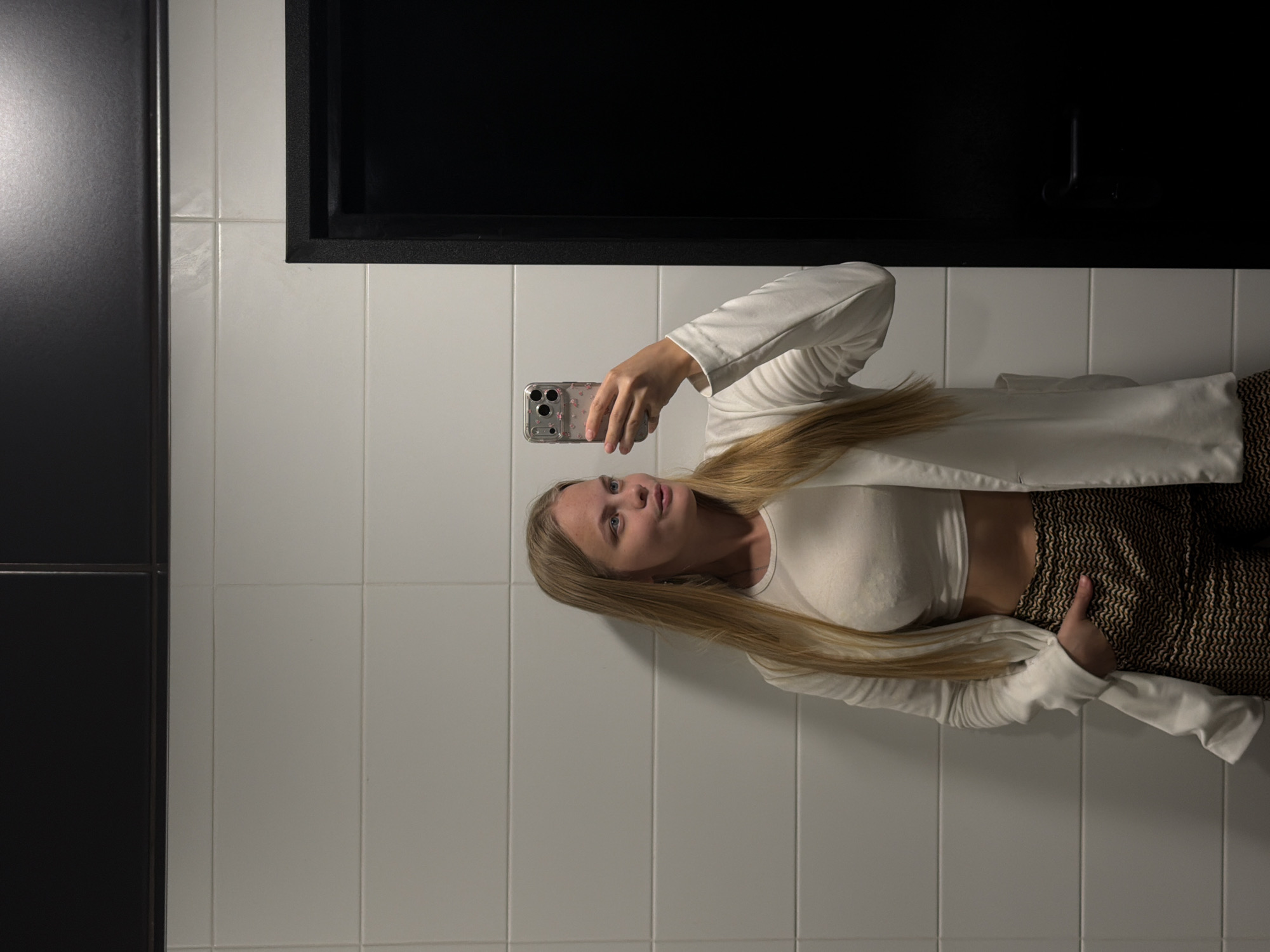 A woman is taking a selfie in a bathroom. The woman has long blonde hair and is wearing a white top and jacket with brown patterned pants. She is holding a cell phone in her left hand, which is adorned with a pink-patterned case. The background consists of white tiled walls and a black storage unit above the woman. The location is likely in Izhevsk, Russia, but the evidence of the location isn't evident in the image.