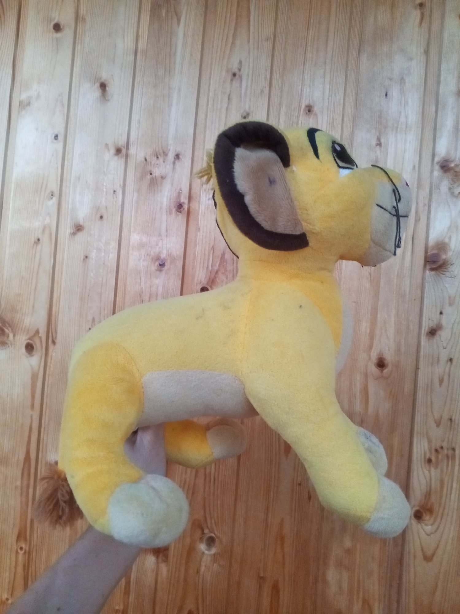 A plush toy of the character Simba from The Lion King is being held up against a wooden paneled wall. The toy is yellow with brown accents on its ears and mane. A human arm is visible supporting the toy from underneath. The image appears to have been taken indoors, likely in a residential setting in Chistopol, Russia, given the subject of the query.