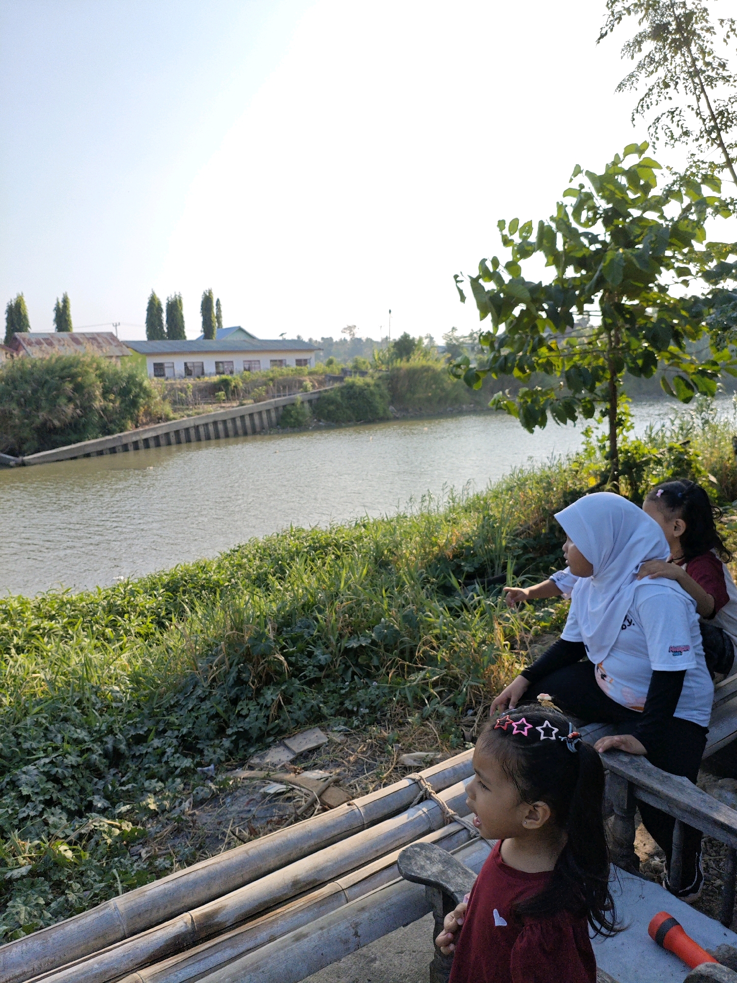 In Padduppa, Indonesia, on a sunny day, three young girls are seen near a body of water. The girl in the foreground, wearing a maroon shirt and a star-shaped headband, looks to her right. Behind her, a woman in a white hijab and a white t-shirt, with a girl in a dark shirt behind her, is pointing towards the water. They are sitting on a wooden structure, likely a bench or platform made of bamboo. The environment includes lush green vegetation lining the water's edge, a calm body of water, and a building with a reddish roof and several windows in the background, possibly a school or administrative building. The scene is illuminated by natural daylight, casting shadows. A red flashlight is visible on the ground to the right of the frame.