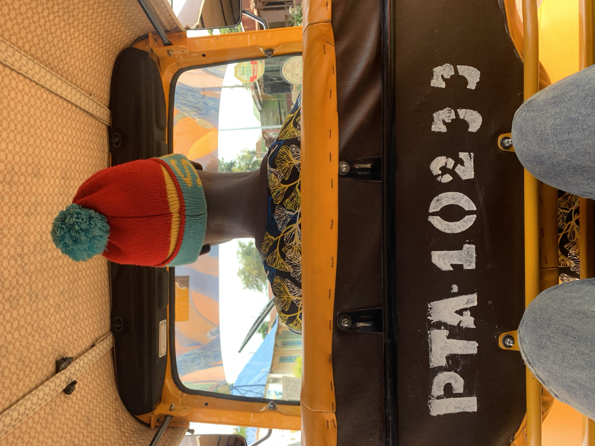 An overhead, forward-facing view from the passenger perspective inside a three-wheeled motor vehicle, likely a keke napep, in New Stadium, Nigeria. The central figure is the driver, seen from the rear, distinguished by dark skin and a prominent red, yellow, and light blue knitted beanie with a teal pom-pom atop his head. He wears a dark-colored shirt featuring a repeated pattern of stylized white and yellow tree-like or floral motifs.
The vehicle's interior roof is covered with a light brown fabric displaying a subtle checkered pattern. Below the ceiling, a dark, curved panel runs across the width of the vehicle. Through the windshield, a semi-urban landscape is visible under bright, possibly overcast, conditions. Buildings with light-colored walls and a distinct blue corrugated metal roof are present in the middle distance, alongside various trees and vegetation. The reflections on the windshield show parts of the interior and distorted external elements. On the driver's side of the windshield, two yellow circular stickers are affixed, one with a green and red emblem, and another with partially visible text, possibly regulatory or identification marks. A QR code is visible on the left side of the windshield.
The back of the driver's seat is upholstered in dark brown material, resembling faux leather. Stenciled in chipped white paint across this surface are the characters "PTA-10233". A yellow padded safety bar runs horizontally across the passenger compartment, positioned between the viewer's legs and the driver's seat. The bottom portion of the image reveals the upper thighs and knees of a passenger, clothed in light blue denim jeans, resting on the seat, confirming the viewpoint.