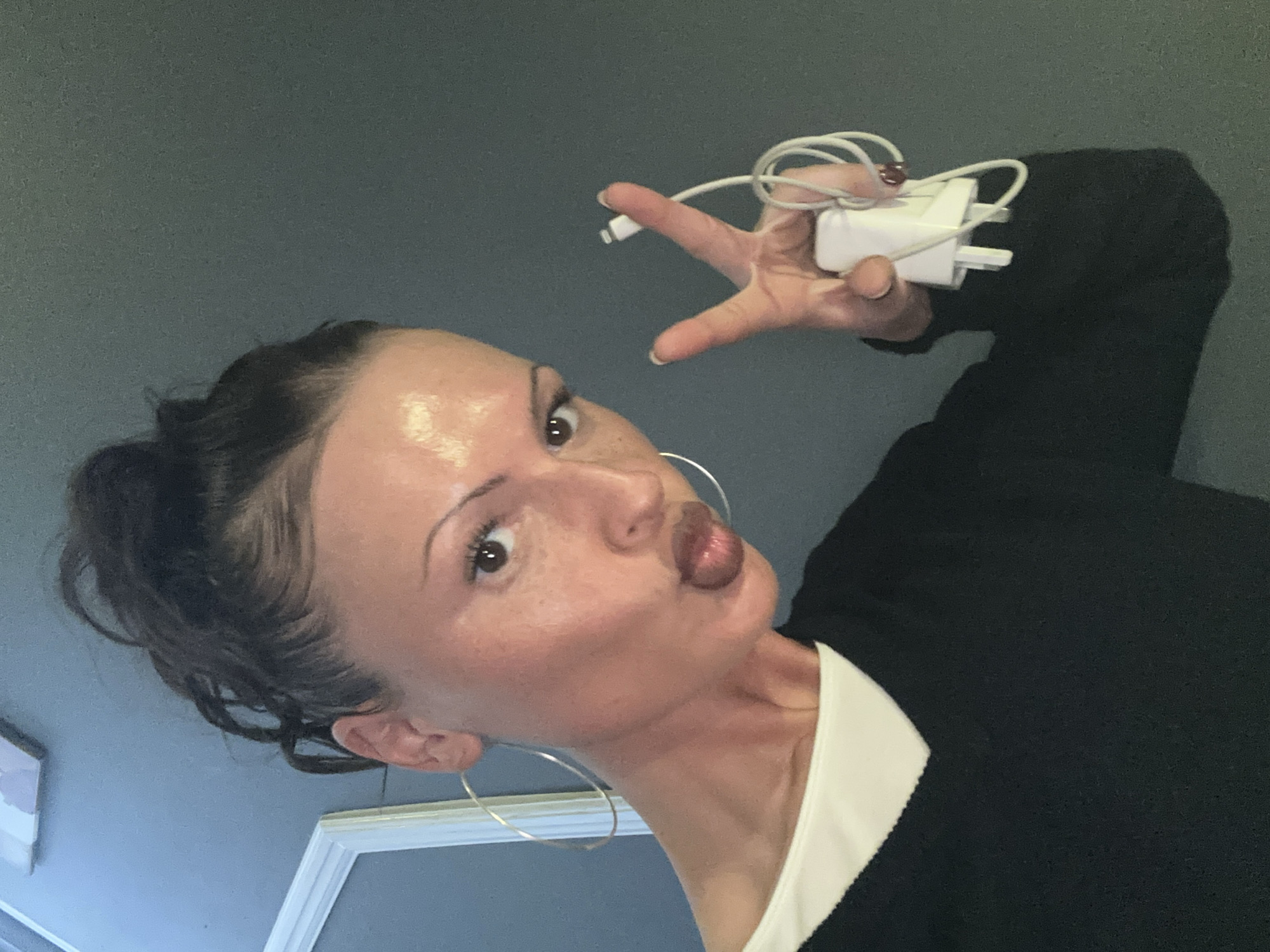 A woman is posing for a selfie. She is holding a white phone charger in her right hand, with the charging cable wrapped around her fingers. Her face is tilted towards the camera, and she is making a duck-lip expression. She has dark hair pulled back into a bun, large hoop earrings, and is wearing a black sweater over a white collared shirt. The background is a gray wall. The location cannot be determined based on the visual information provided.