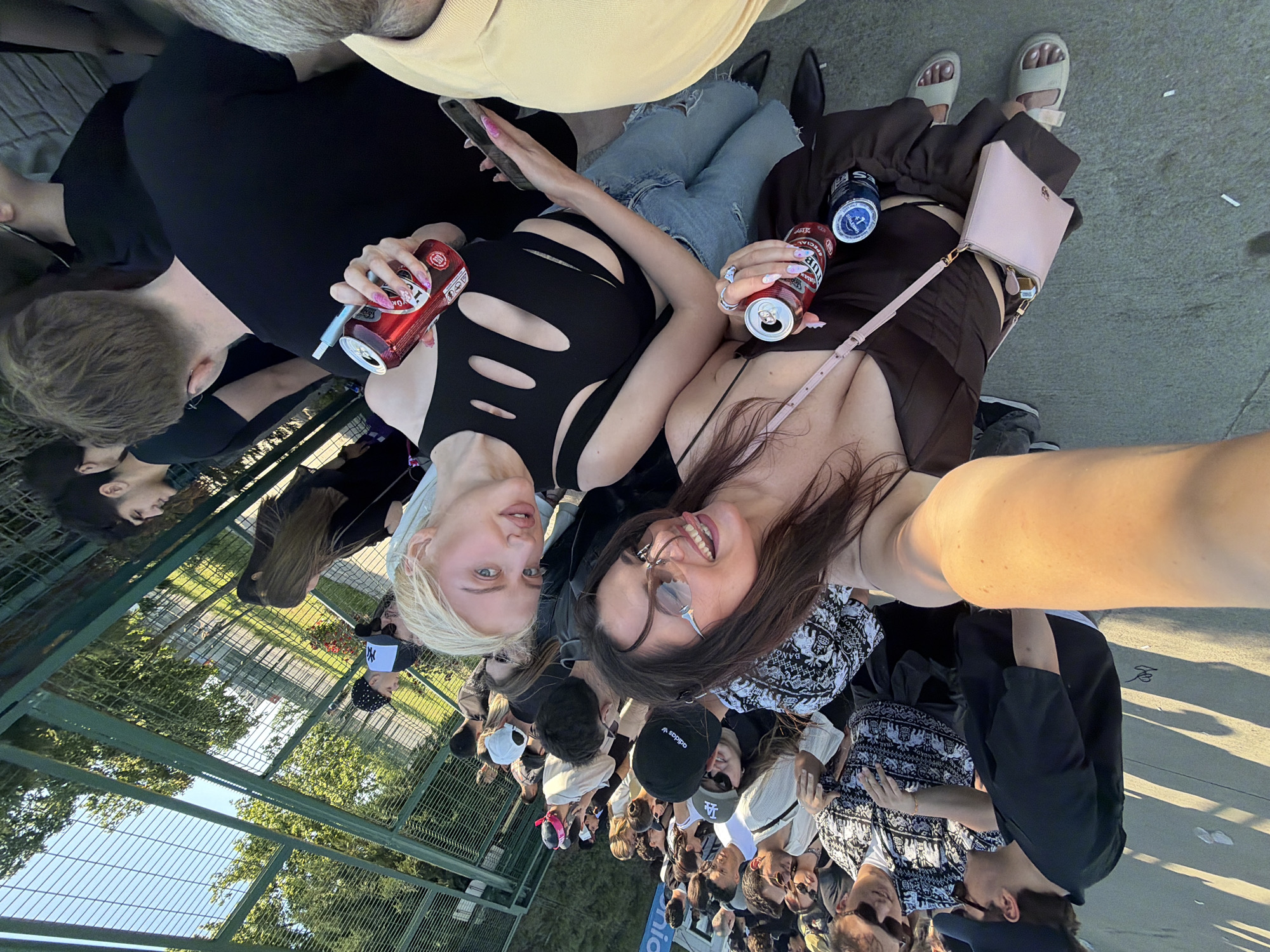 A slightly rotated outdoor selfie captures two young women in the foreground, each holding an opened can of PUB beer. The woman on the left has blonde hair and wears a black cut-out top, paired with ripped blue jeans. The woman on the right has long dark hair, glasses, and a brown top, with a small pink purse at her side. Both are facing the camera and smiling.
Behind them, a large crowd of people extends into the background. A green chain-link fence and trees are visible in the upper left corner of the frame.
The location is suggested to be Başakşehir, Turkey. A concrete surface serves as the ground beneath the subjects.