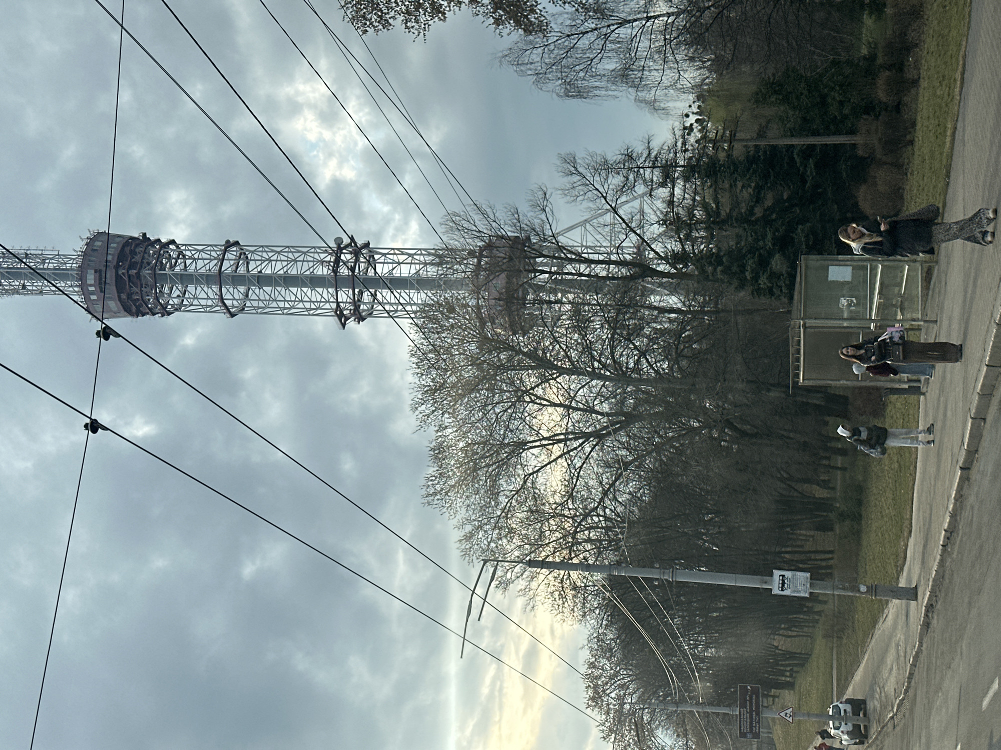 The image captures the Kyiv TV Tower in Kyiv, Ukraine. The tower is a tall, lattice-structured metal tower that dominates the scene. The foreground shows people walking along a sidewalk. The background is a cloudy sky and bare trees. Power lines are visible in the foreground, extending from the street up towards the tower. The lighting suggests it is daytime, but the sky is overcast. There's a bus stop on the side of the road. The area appears to be a park setting.