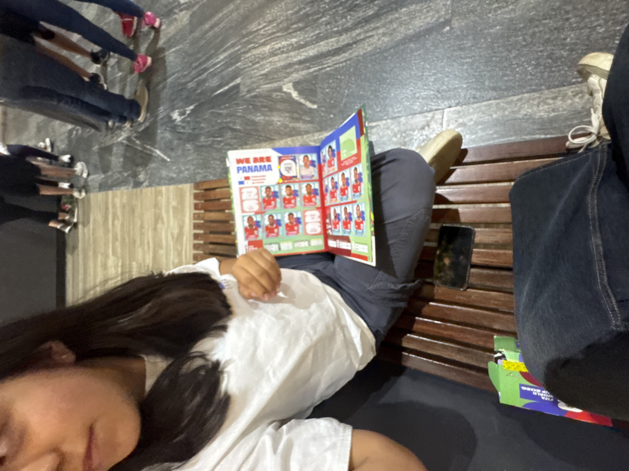 A person with long dark hair is lying on a brown slat bench, reading a book titled "WE ARE PANAMA". The person is wearing a white shirt and gray pants. A black smartphone is on the bench next to them. Adjacent to them sits another person wearing jeans and light colored sneakers. On the bench between the two people is what appears to be a box with "FIFA WORLD CUP 2026" printed on it. Several other people can be seen in the background in the distance. The ground and nearby wall have a marble like pattern. No details visible indicate a specific location in Puebla, Mexico.