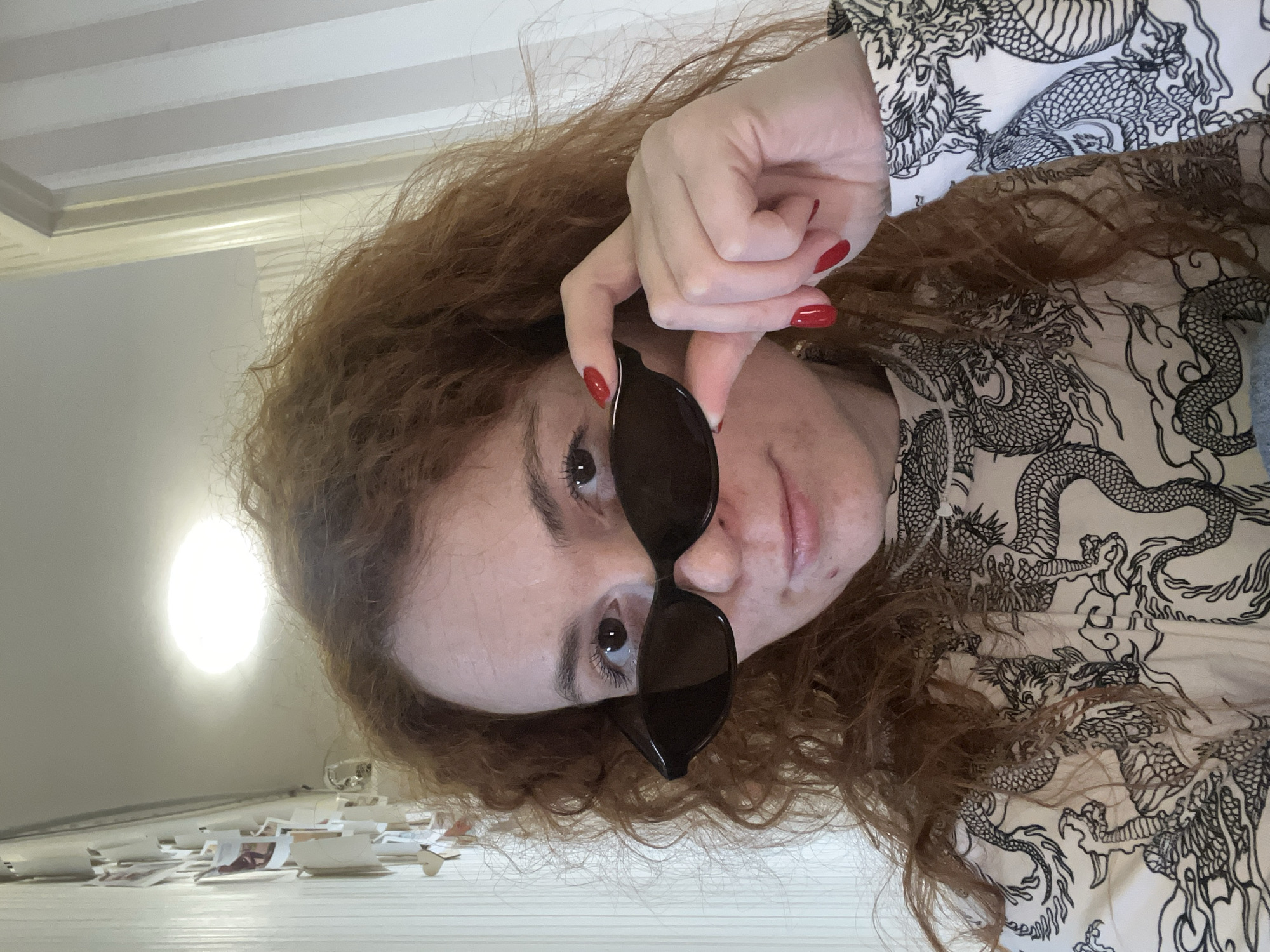 An eye-level selfie shows a person with long, wavy reddish-brown hair, tilted 90 degrees clockwise, making the person's head appear on the left side of the frame. The person is holding a pair of black sunglasses, with their right hand visible, featuring bright red nail polish on the fingernails. One eye is partially visible above the sunglasses, looking directly at the camera with a neutral expression, revealing dark eyelashes and a light brown iris. The person wears a light-colored shirt adorned with intricate black dragon patterns.
The background suggests an indoor setting. Above the person's head (which is to the right in the rotated image), a ceiling with decorative white molding is visible. To the lower left (which would be below the person's feet in a normal orientation), a bright, circular light source illuminates the scene. Below this light, on a light-colored surface, several small, scattered pieces of paper or similar light-colored debris are visible. No text is visible in the image.