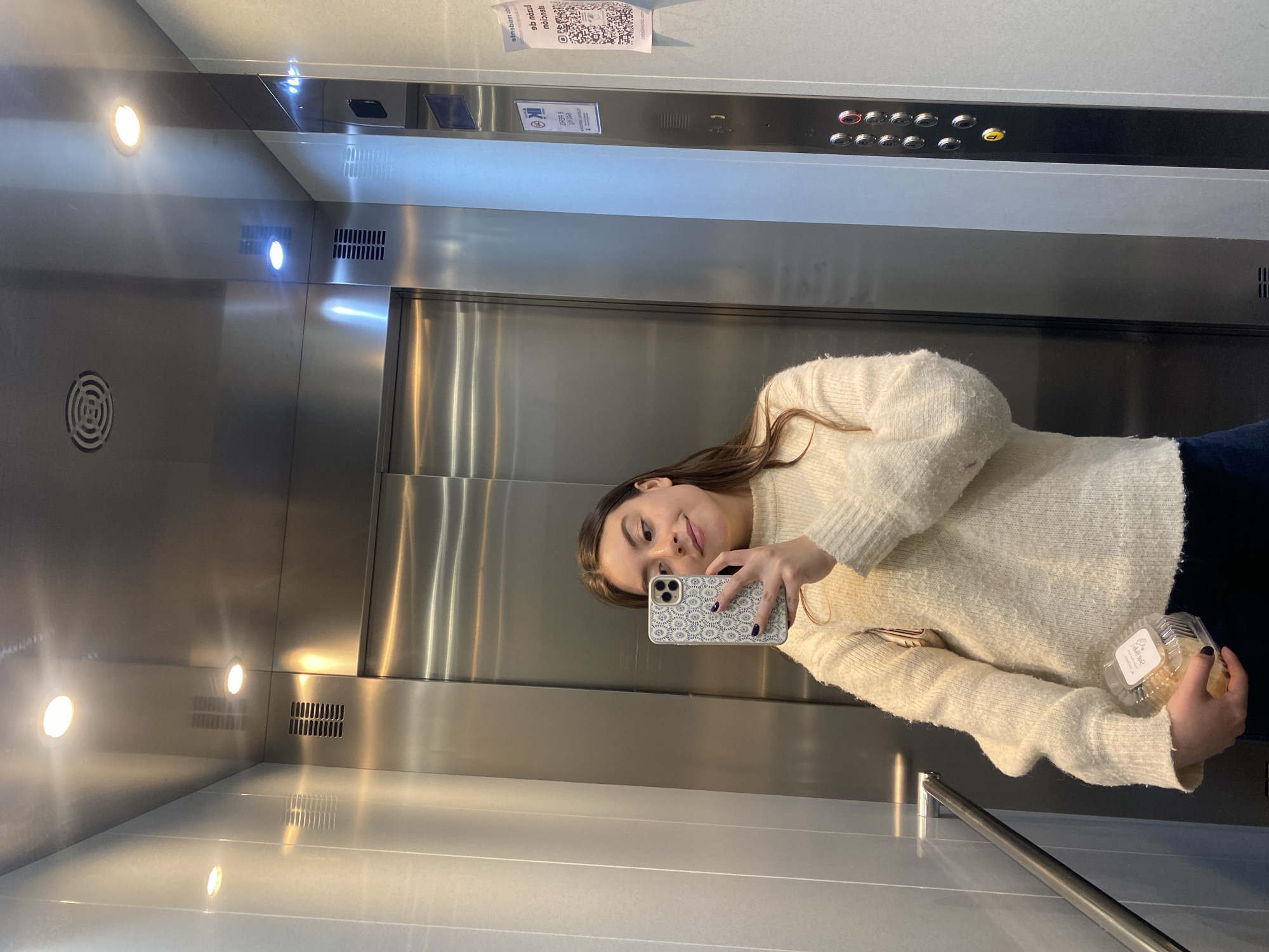 A woman is taking a selfie in an elevator. She is holding a smartphone in her left hand and a small clear plastic container in her right hand. The elevator has a stainless steel interior with buttons, lights, ventilation grilles, and signage. The location is Unknown, Unknown.