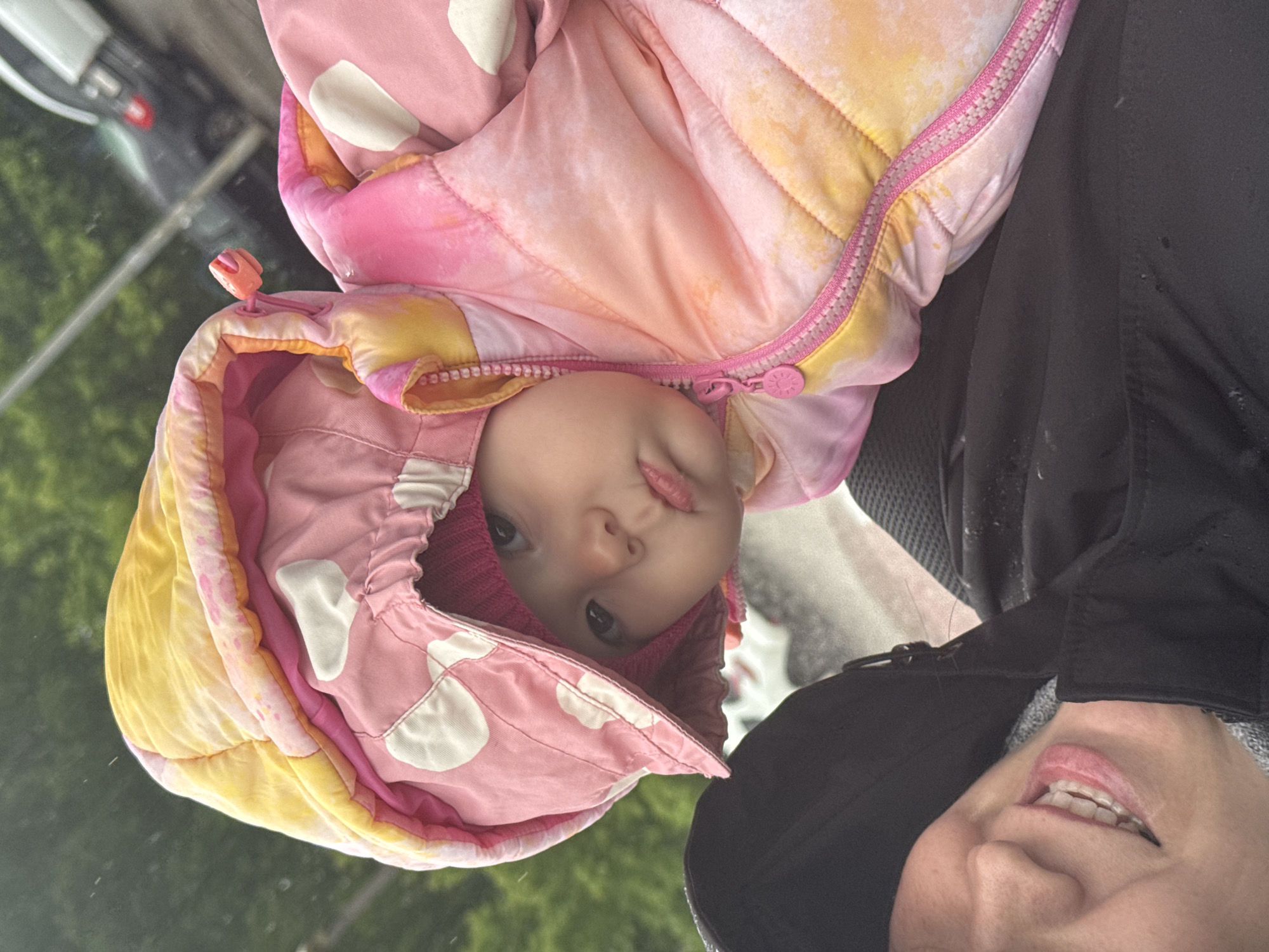 A close-up, canted shot shows a baby in a pink and yellow polka dot puffy snowsuit. The baby's face is centered, peering out from the hood of the suit. A woman's face is visible in the lower right, indicating she is holding the baby. The background suggests an outdoor setting, possibly with vehicles visible through a reflective surface. The location, Zav'yalovo, Russia, cannot be confirmed based on the image.
