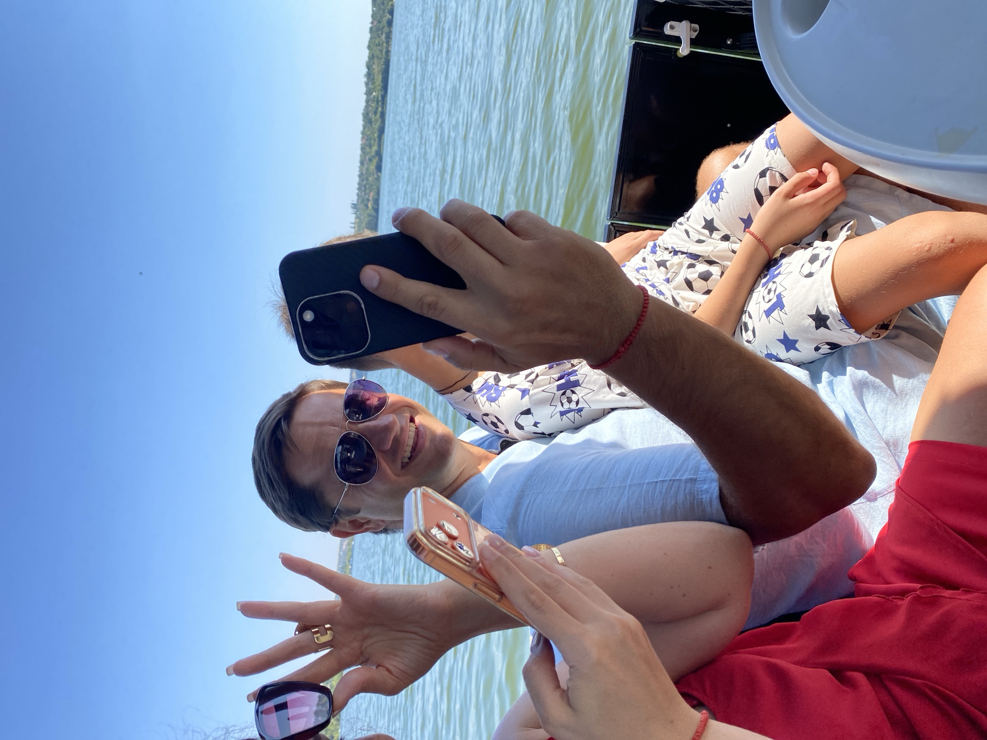 The image depicts a group of people on a boat, with a clear blue sky and a body of water in the background. The perspective suggests the photo is taken from within the boat, looking towards the bow. In the foreground, a woman's hand, adorned with rings, holds up sunglasses. To her right, a man wearing sunglasses and a light blue shirt smiles at the camera, holding up a smartphone to take a selfie. Another person, a child wearing shorts with a soccer ball pattern, is seated next to the man, their arm resting on the man's leg. The child is also wearing a red bracelet. The overall scene suggests a casual, outdoor gathering on a sunny day. The location is identified as Molovata, Moldova, implying the boat is on the Nistru River near the village.