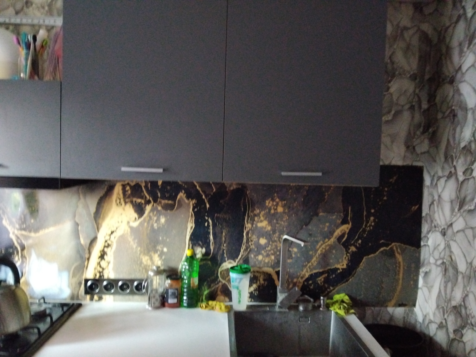 The image shows a kitchen scene. The upper part of the image features two dark gray cabinets with minimalist handles. Below the cabinets is a backsplash with a marbled pattern in black, gold, and white. To the left, a stovetop with a silver kettle is visible. In the center, a stainless steel sink with a silver faucet is present. On the countertop next to the sink are several items, including a green bottle, a jar, and a shaker cup. To the right of the sink, a dark object, possibly a trash can, is partially visible. The right side of the image shows a wall with a similar marbled wallpaper pattern. The scene depicts a domestic kitchen environment.