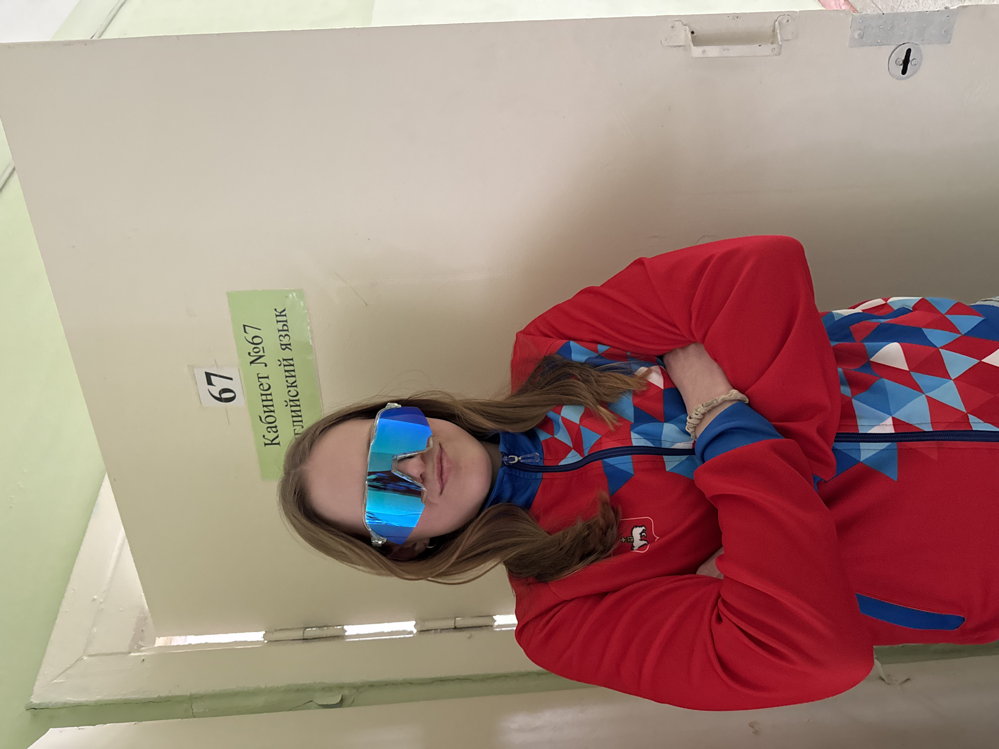 A person is standing against a light-colored door in what appears to be a room or hallway. The door has a sign in Russian that includes the number "67". The person is wearing sunglasses that reflect the surroundings and a red jacket with blue and white geometric patterns. A logo is visible on the jacket. The location is Usolye, Russia.
