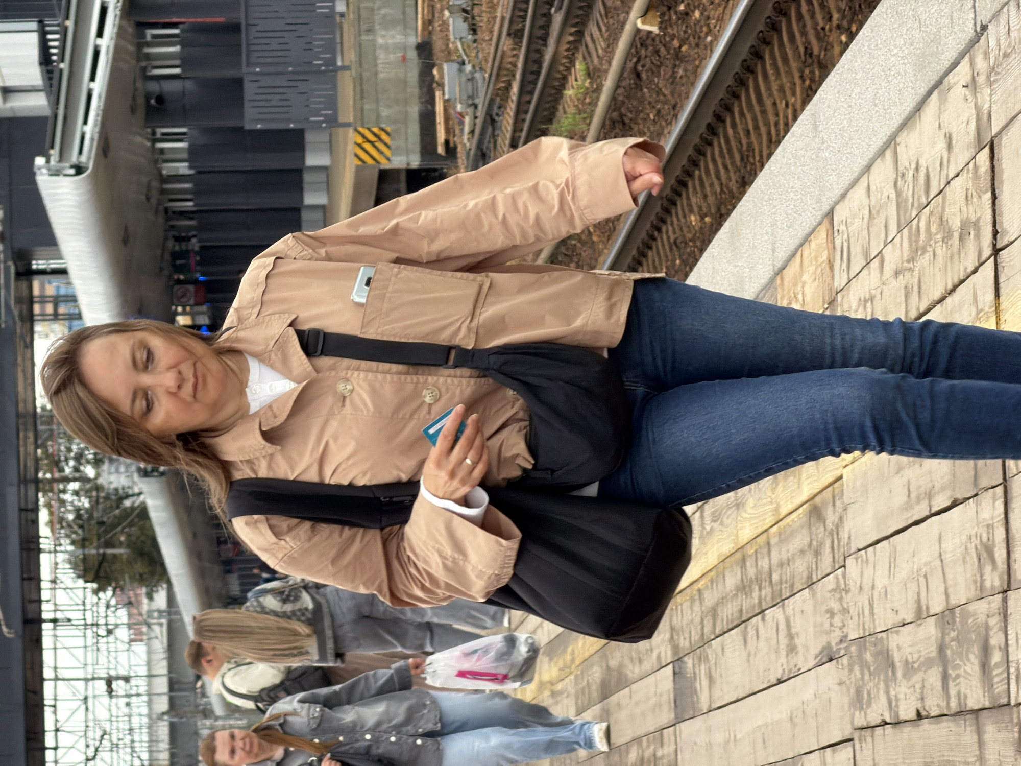 An adult woman with light brown hair, wearing a light brown jacket over a white shirt and blue jeans, walks on a wooden platform next to train tracks. She carries a black bag slung across her body and holds a blue card in her right hand. A silver smartphone is visible in the chest pocket of her jacket. In the background, other individuals are present on the platform, and a covered structure with pillars is visible. The environment appears to be a train station platform.