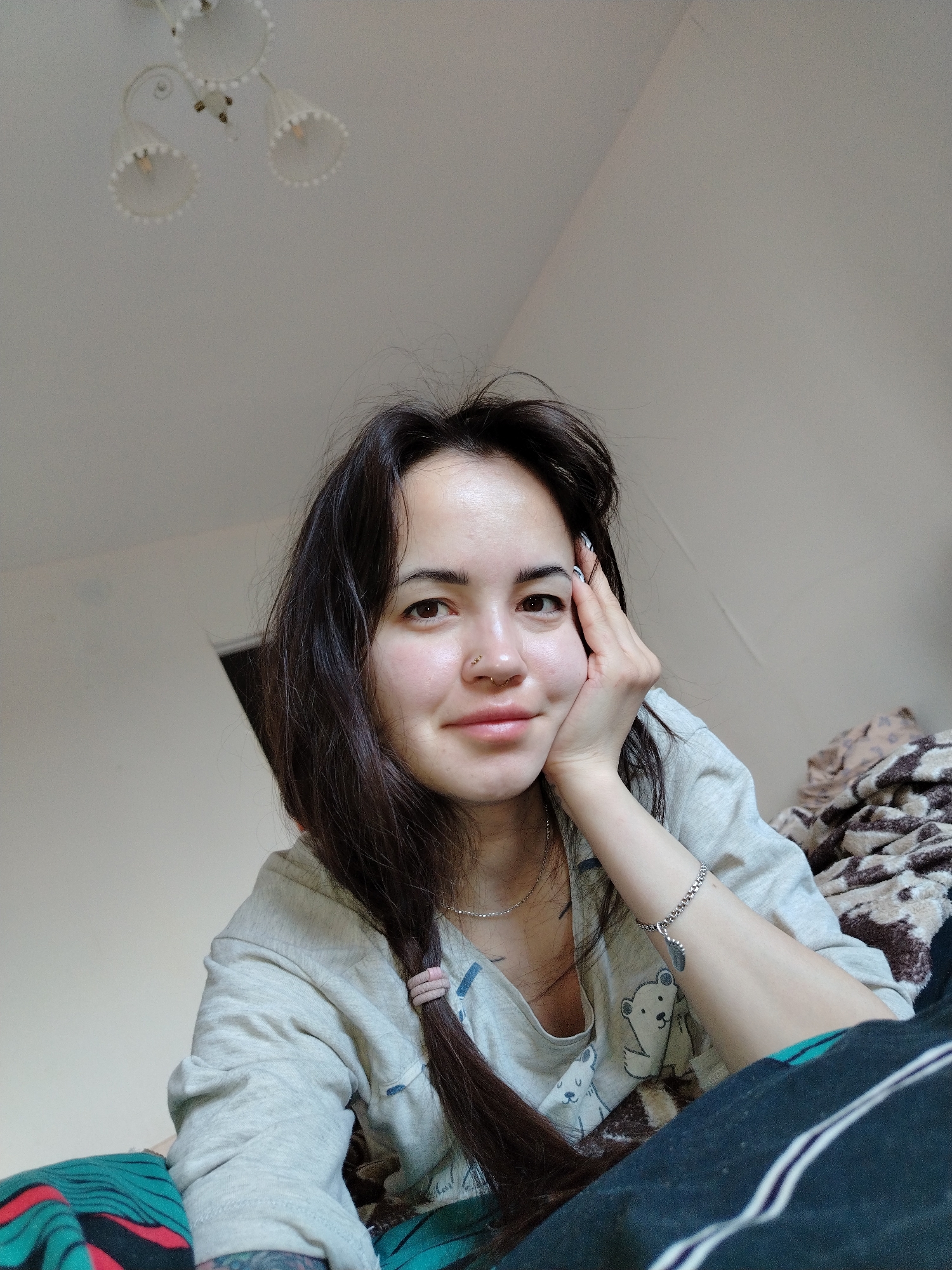 A young woman with long, dark hair, fair skin, and nose piercings (septum and nostril) is shown in a relaxed, close-up indoor portrait. She looks directly at the camera with a gentle, contented smile, her left hand lightly resting against her cheek. She is wearing a light grey long-sleeved top that features a graphic of a polar bear on the front. A silver chain necklace and a silver bracelet with a footprint charm are visible accessories. Her hair is casually styled, with a section braided and secured by a pink elastic.
The setting appears to be a cozy indoor space, likely a bedroom or living room in Noviy, Russia, suggested by the casual attire and visible bedding. In the upper left background, a white ceiling chandelier with three light fixtures is visible. The walls are a plain, light neutral color. Surrounding the woman are various patterned blankets or bedding in shades of teal, red, black, beige, and brown. The overall atmosphere conveyed is calm and comfortable.