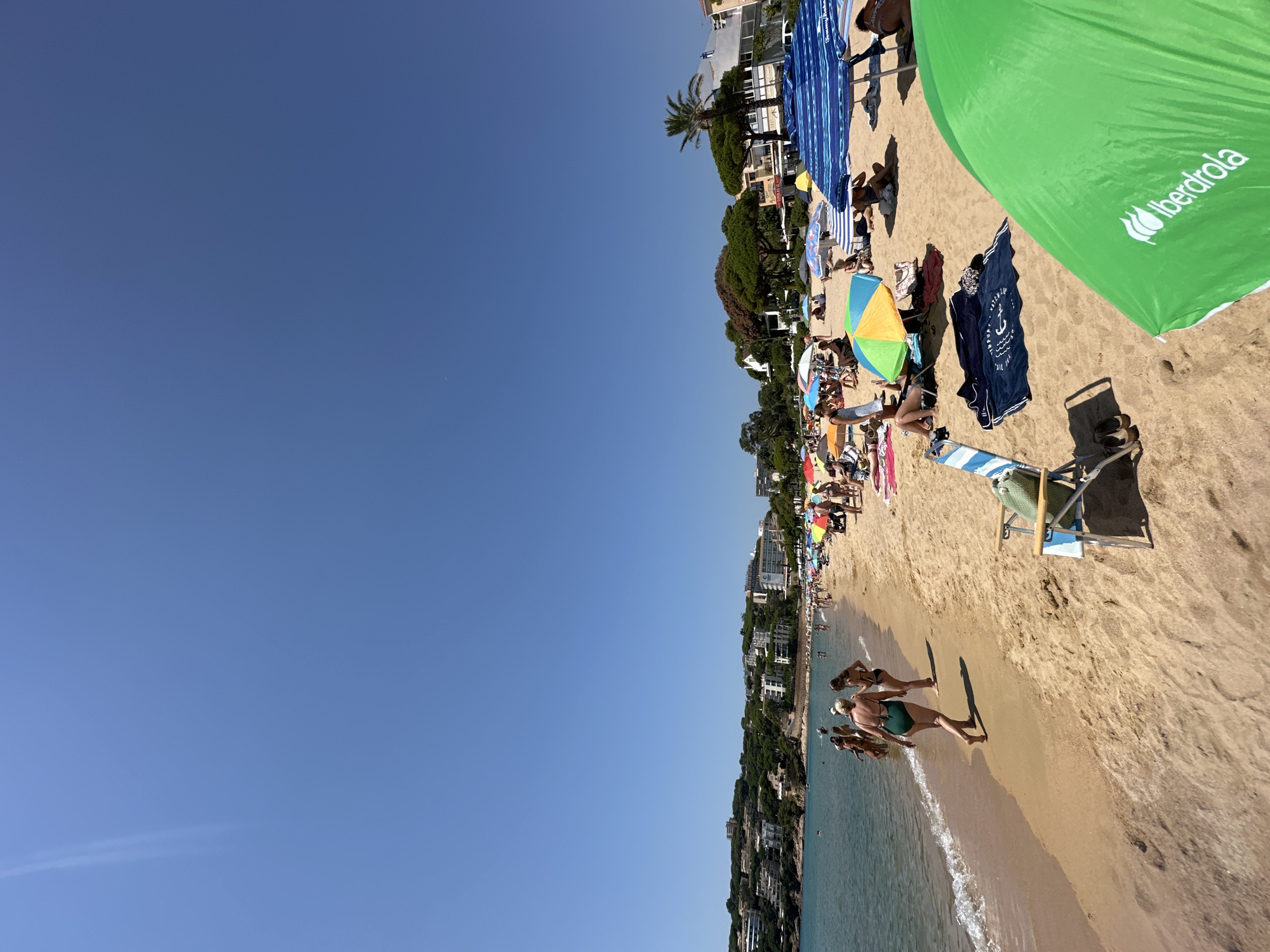 The image captures a crowded beach scene in Sant Feliu de Guíxols, Spain, on a clear, sunny day. The beach is sandy, with a gentle slope towards the calm, blue water. Numerous people are present, engaged in various beach activities. Several individuals are walking along the shoreline, while others are seated or lying on towels and beach chairs under a variety of colorful umbrellas, including large blue and white striped ones and smaller, vibrant multi-colored ones. Some people are in the water. In the background, the coastline is lined with lush green trees and densely packed buildings, characteristic of a coastal resort town. The sky is a bright, cloudless blue. A prominent green umbrella with white text, likely a brand logo, is in the foreground on the right side of the frame. The overall atmosphere is one of leisure and summer enjoyment.