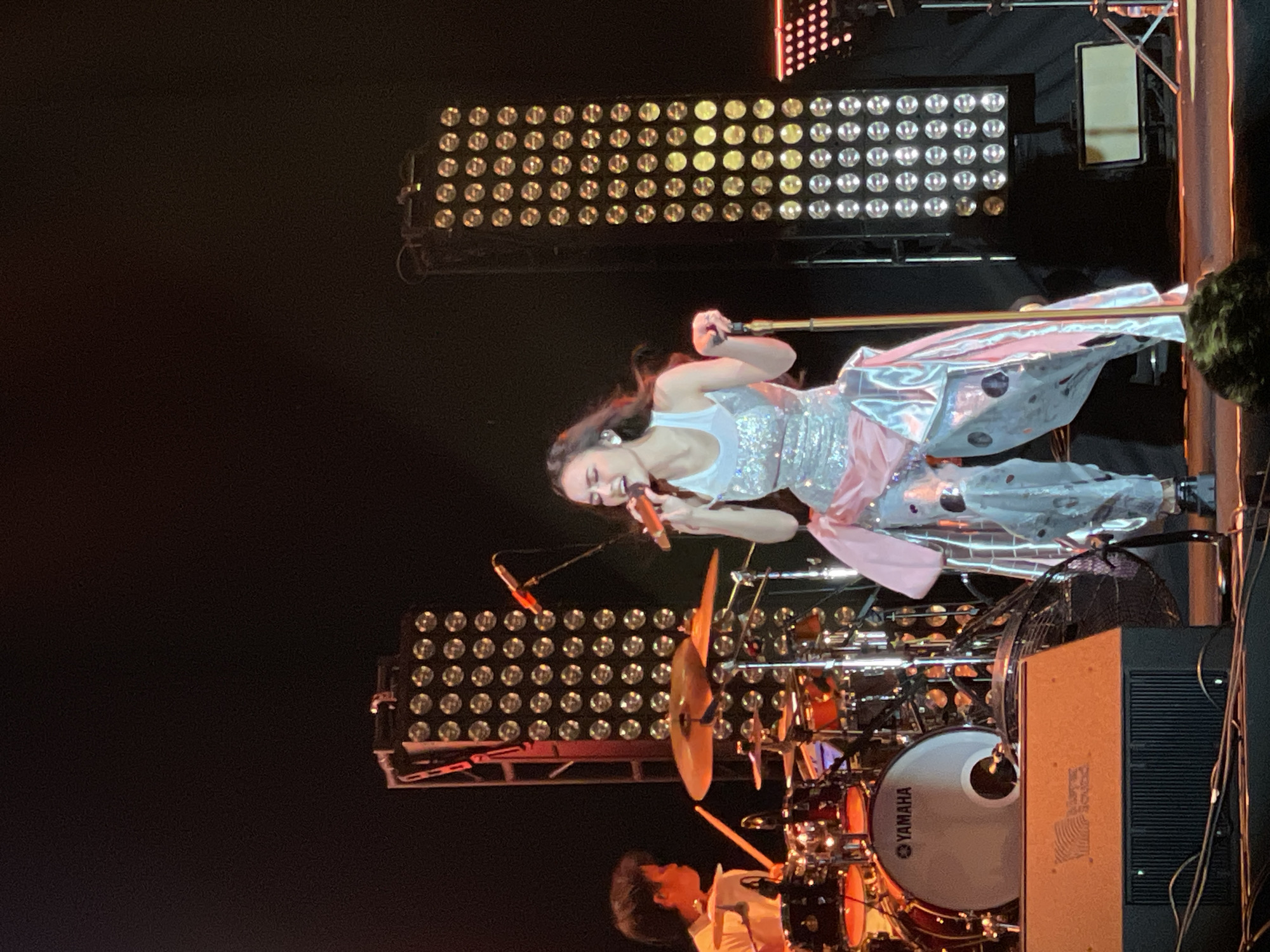 A female performer is on stage at an indoor venue in Salembaran, Indonesia. She is wearing a light-colored sequined top and metallic pants with a pink sash, singing into an orange microphone. Behind her are two large light arrays and a drum set with a Yamaha branded bass drum. A drummer is visible behind the kit.
