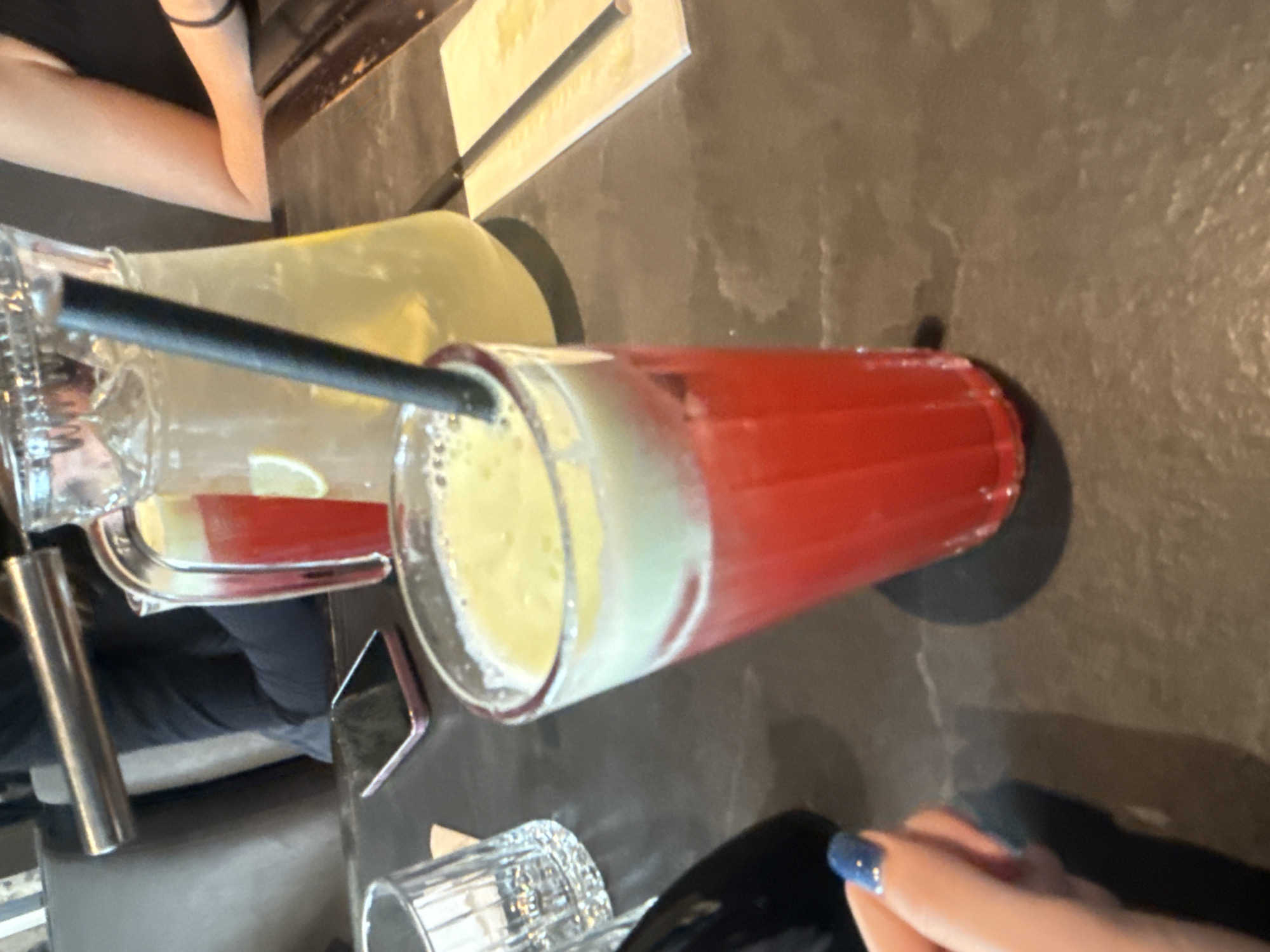 The image shows a close-up of drinks on a dark-colored table. A glass containing a red-colored beverage with a yellow, frothy layer on top sits next to a pitcher of yellow liquid. A black straw is inserted into the glass. The table has a textured surface. A hand with blue nail polish is visible in the foreground. In the background, there are partial views of people and a metal object. The location is not identifiable as Saint Petersburg, Russia, based solely on the image.