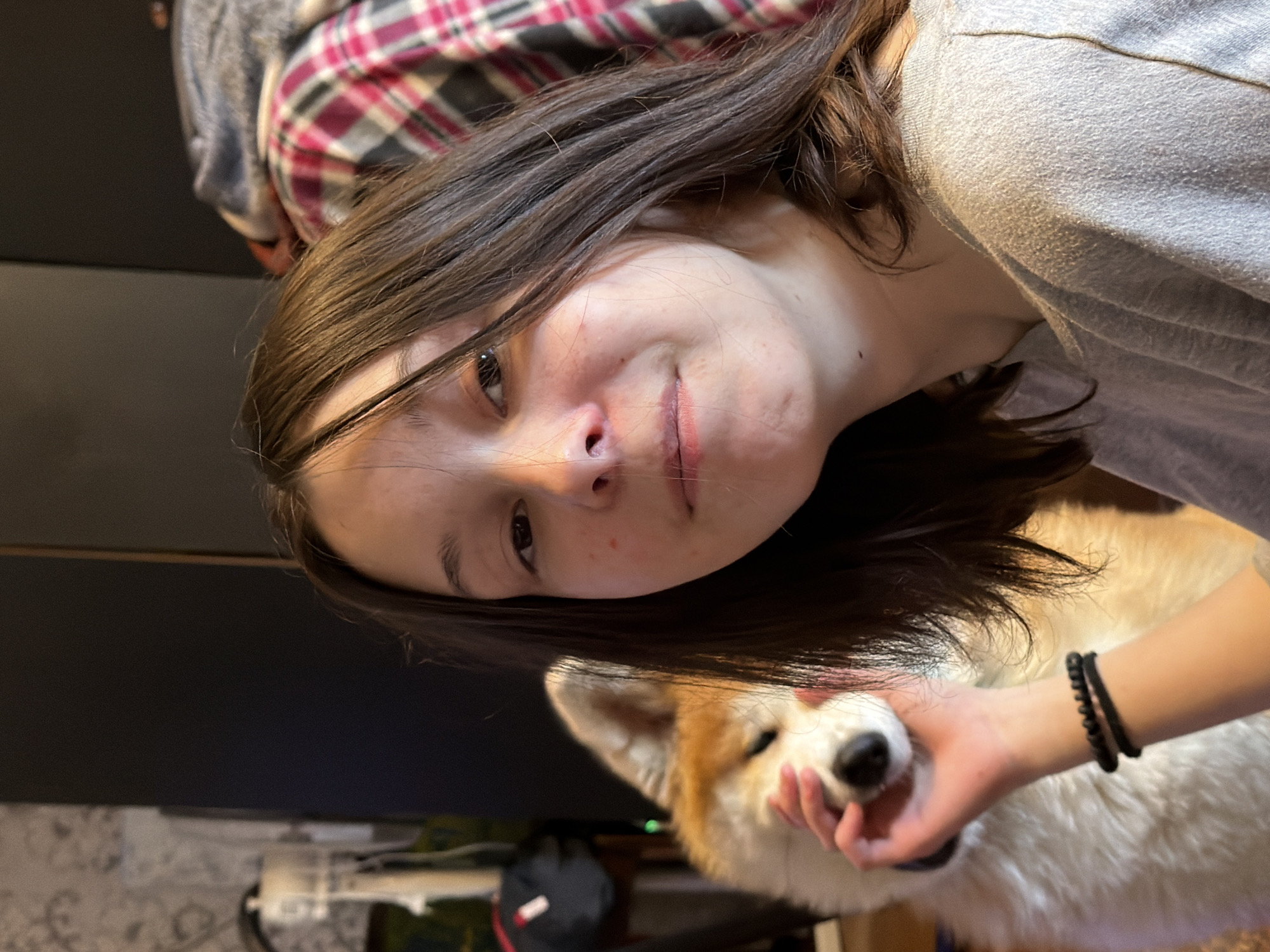 A young woman with dark hair is holding a white and tan dog, possibly an Akita, with her right hand. She is wearing a gray long-sleeved shirt and has a black beaded bracelet on her right wrist. The dog is facing the camera. The background includes a dark-colored wall, a plaid shirt, and what appears to be a room with a patterned floor and electronic equipment. There is no visual evidence to support the location being Заболотово, Russia.