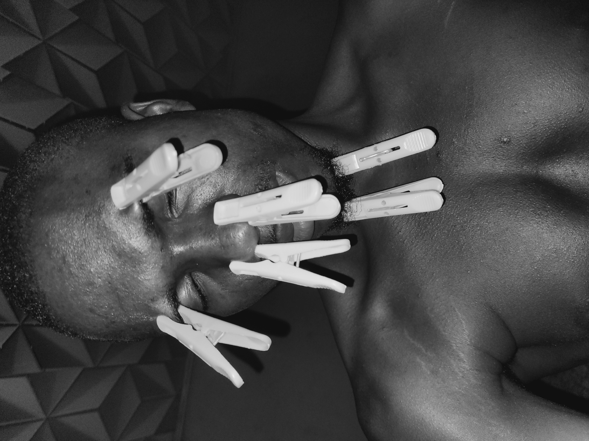 The black and white image shows a close-up of a Black man's face and upper chest. The man's eyes are closed, and he has several white clothespins attached to his face, including on his eyelids, nose, and chin. The background consists of a textured surface with a geometric pattern. The man's skin appears to be sweating. The image is a portrait with the man as the central subject.
