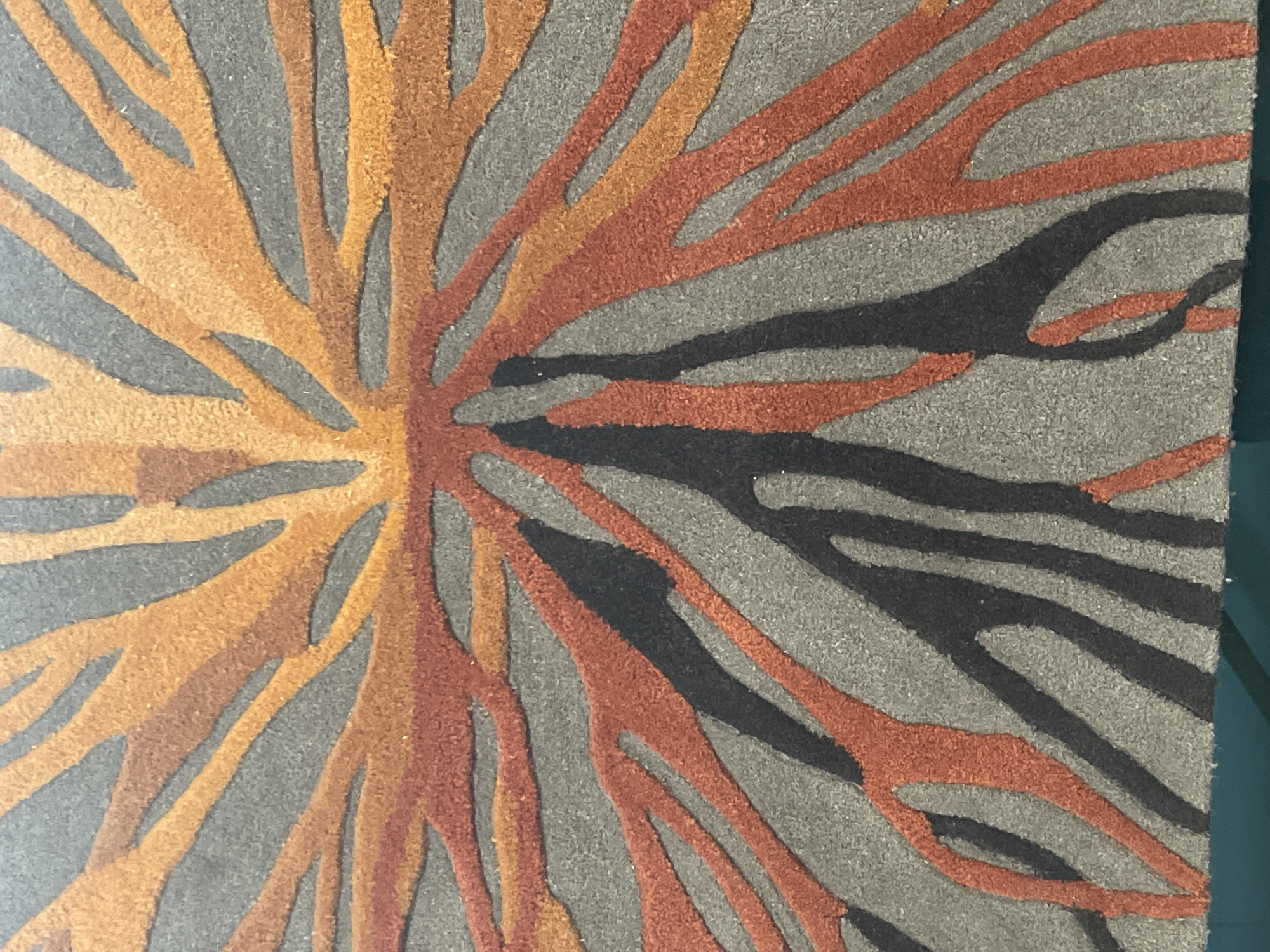 The image shows a close-up, overhead view of a textured rug with an abstract design. The design features radiating lines of various earthy tones, including orange, rust, and dark brown or black, set against a neutral gray background. The lines are thick and organic, resembling tree roots spreading outwards. The rug's texture appears to be made of short, dense fibers, giving it a slightly fuzzy appearance. The lighting is even, with no harsh shadows, suggesting an indoor setting. The scene is static, with no people or discernible actions. The location is unknown.