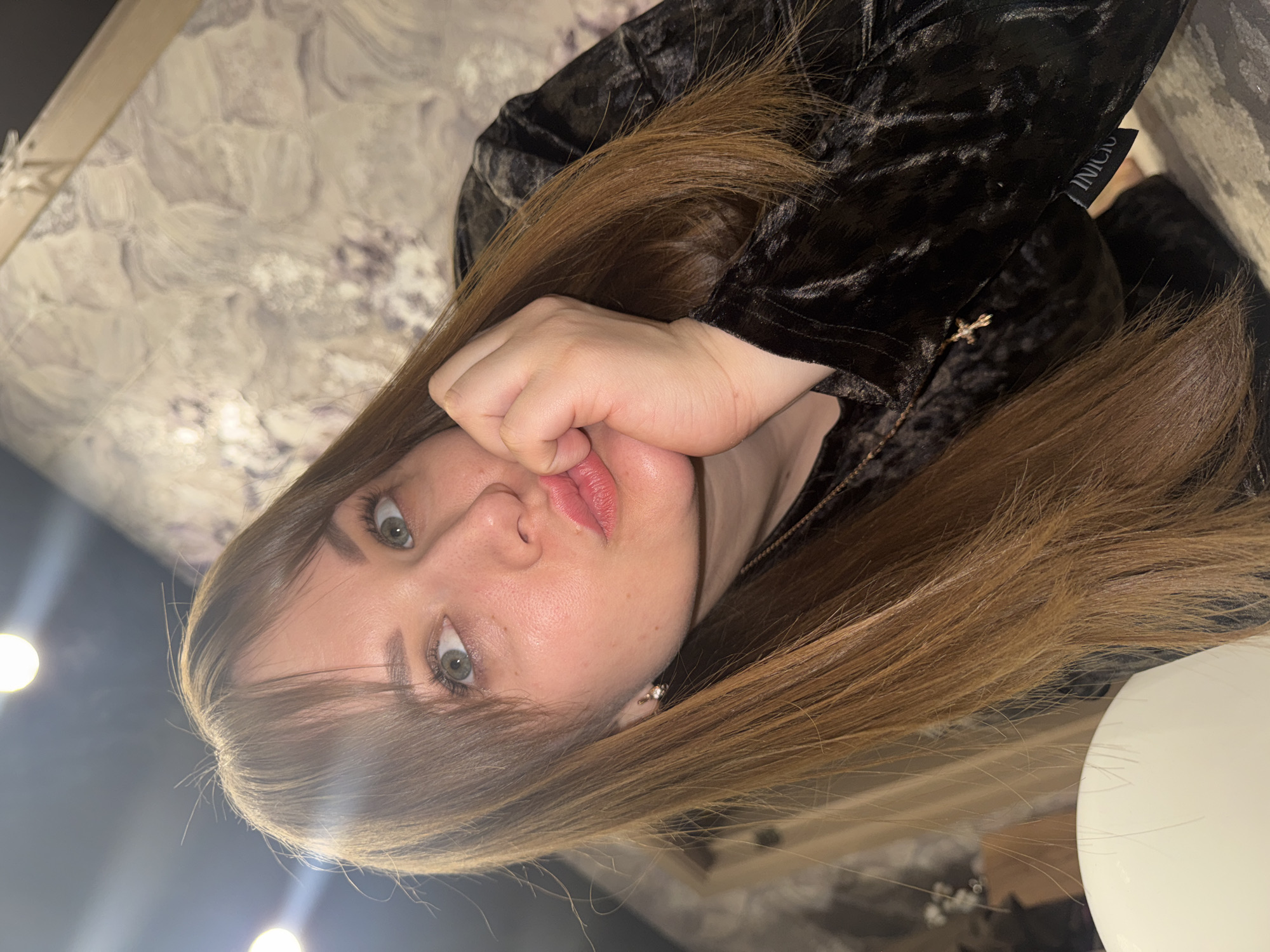 A rotated, close-up portrait features a young woman with long, straight, light brown hair and bangs, looking directly at the viewer. Her eyes are a light green or hazel, and her lips are subtly parted, appearing to have light pink lipstick. She has fair skin.
Her left hand is positioned near her face, with her thumb tucked under her chin and her fingers touching her lips, conveying a pensive or slightly posed expression. She is wearing a dark, possibly black, velvet-textured garment, which appears to be a jacket or top. A small, gold-colored cross charm is visible on a chain near her collarbone. On a black tag attached to her clothing, the word "INICIO" is visible in white text.
The background consists of a patterned wall with a light gray and white, marble-like or swirly design. Several blurry, circular light sources, possibly ceiling lights or lamps, are visible, indicating an indoor setting with artificial lighting, likely suggesting evening or night. A portion of a white spherical object is visible on the right side of the frame. The image offers no specific visual cues to identify its location, beyond the provided context of Chaikovsky, Russia.