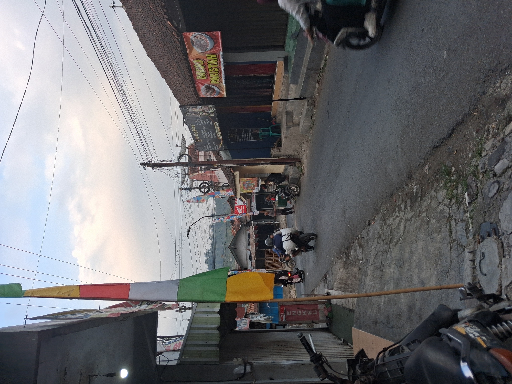 The image depicts a street scene in Bantarkawung, Indonesia. A prominent multi-colored flag, composed of green, yellow, red, and white sections, hangs from a wooden pole in the foreground on the left. Adjacent to the flag is a building with a gray facade and a metal shutter. Across the asphalt road, several buildings are visible, including one with a brown tiled roof and a banner that reads "BAKSO PAKISTAN." Several people are on motorcycles, with two individuals on one motorcycle in the center of the frame transporting large white sacks. Another person is seen walking on the sidewalk on the right side of the road. The sky is overcast with scattered clouds. The environment appears to be a commercial or residential street with a utilitarian aesthetic.