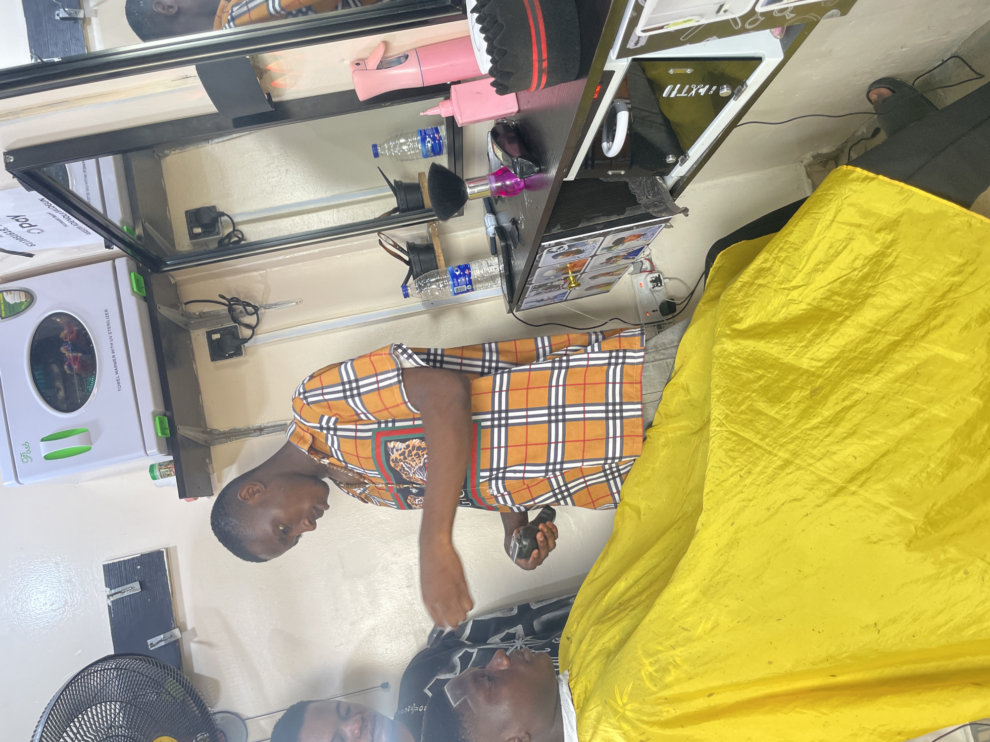 Inside a barber shop in Ado, Nigeria, a barber in an orange and white plaid shirt gives a haircut to a client under a yellow cape. On a shelf behind the barber are clippers, water bottles, and other barbering tools. A fan is visible in the lower left corner of the frame. The barber and client are both dark-skinned.