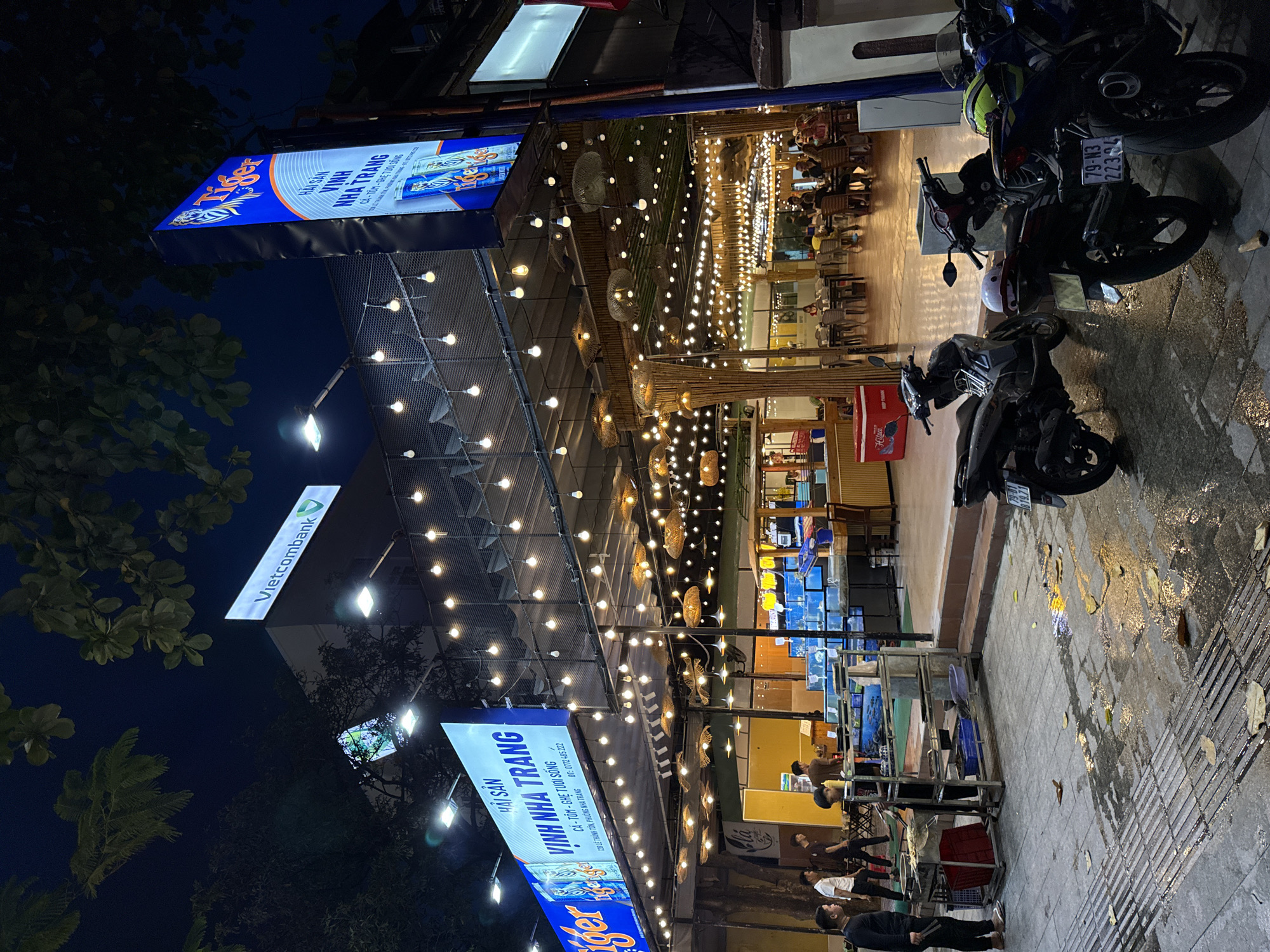 The image depicts a nighttime street scene in Nha Trang, Vietnam, focusing on a seafood restaurant called "HẢI SẢN VINH NHA TRANG". The restaurant is brightly lit with numerous string lights and spotlights illuminating the outdoor dining area and the seafood tanks. The facade features a large blue sign with "Tiger" branding and text that translates to "Seafood Vinh Nha Trang". Inside the restaurant, tables and chairs are set up for patrons, and some people are visible dining. Outside, a food preparation area with a metal cart is set up, and a few men are standing nearby, possibly employees. Several motorcycles are parked on the wet pavement in front of the establishment, indicating the common mode of transportation in the area. The overall atmosphere suggests a lively and active evening at a popular local eatery.