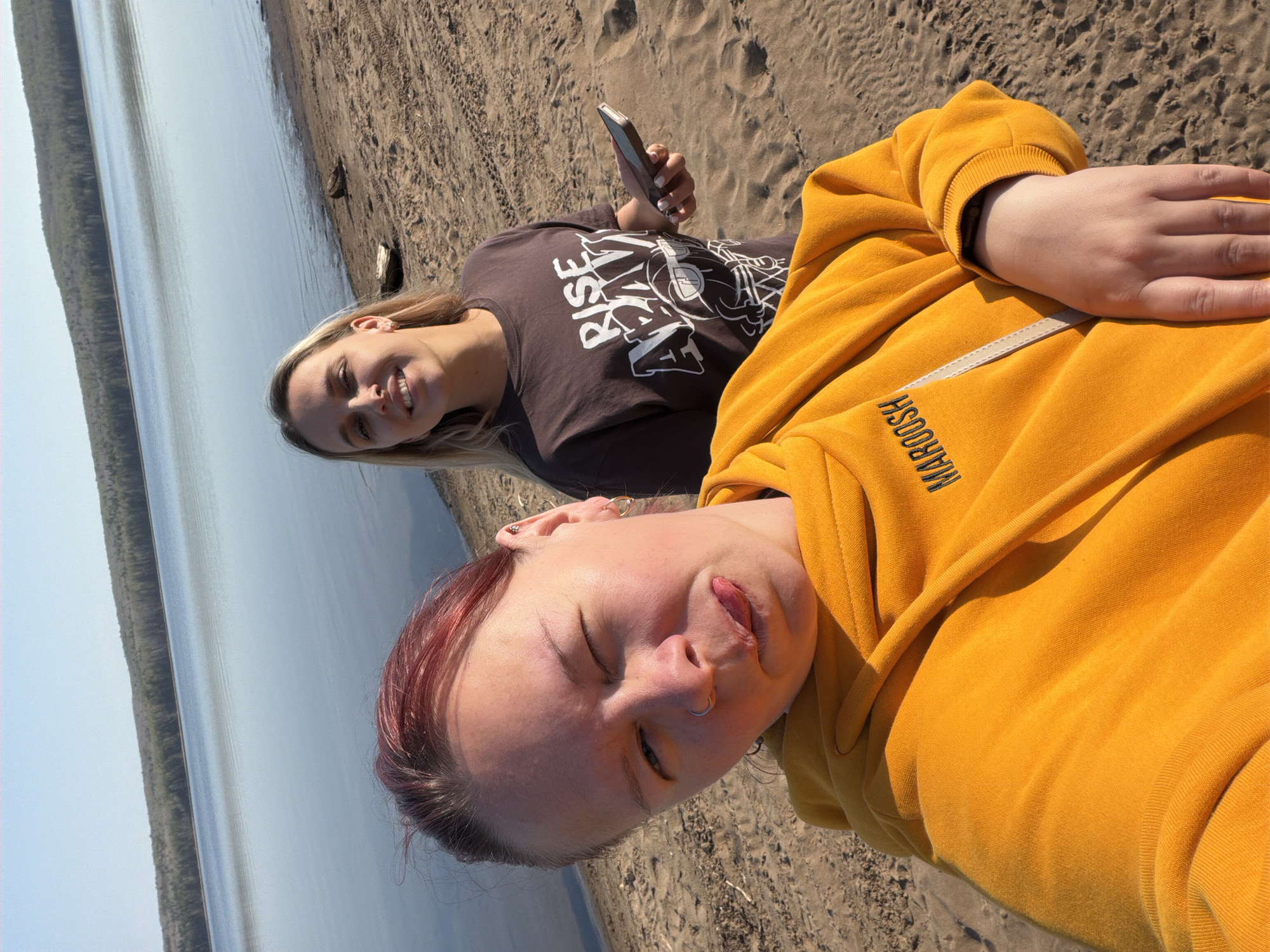 Two women are on a sandy beach, with a body of water and trees in the background. The woman in the foreground is wearing a yellow hoodie and has red-tinted hair; she is sticking her tongue out and has her eyes mostly closed. The other woman, further back, is holding a smartphone and is wearing a brown t-shirt with the word "RISE" on it. The location is Chaikovsky, Russia.