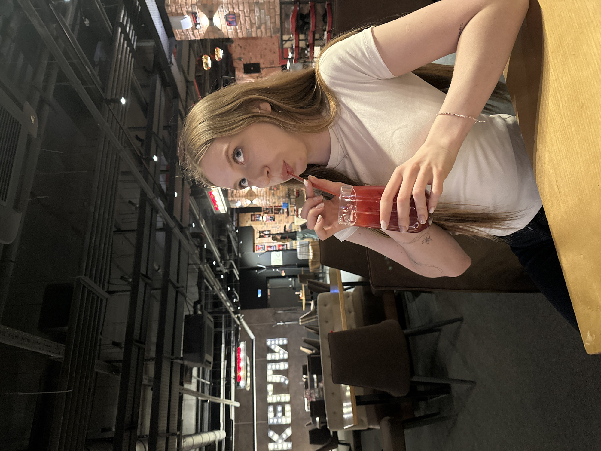A woman with long blonde hair wearing a white t-shirt is seated at a table and drinking from a glass filled with a red liquid through a straw. She is holding the glass with her left hand. The location appears to be a restaurant or cafe in Izhevsk, Russia, as the sign "КЕЙСТИ" (KEJSTI) is visible in the background, which is a known establishment in that city. The background shows a busy interior with tables, chairs, and industrial-style overhead structures. The lighting is dim, creating a casual atmosphere.
