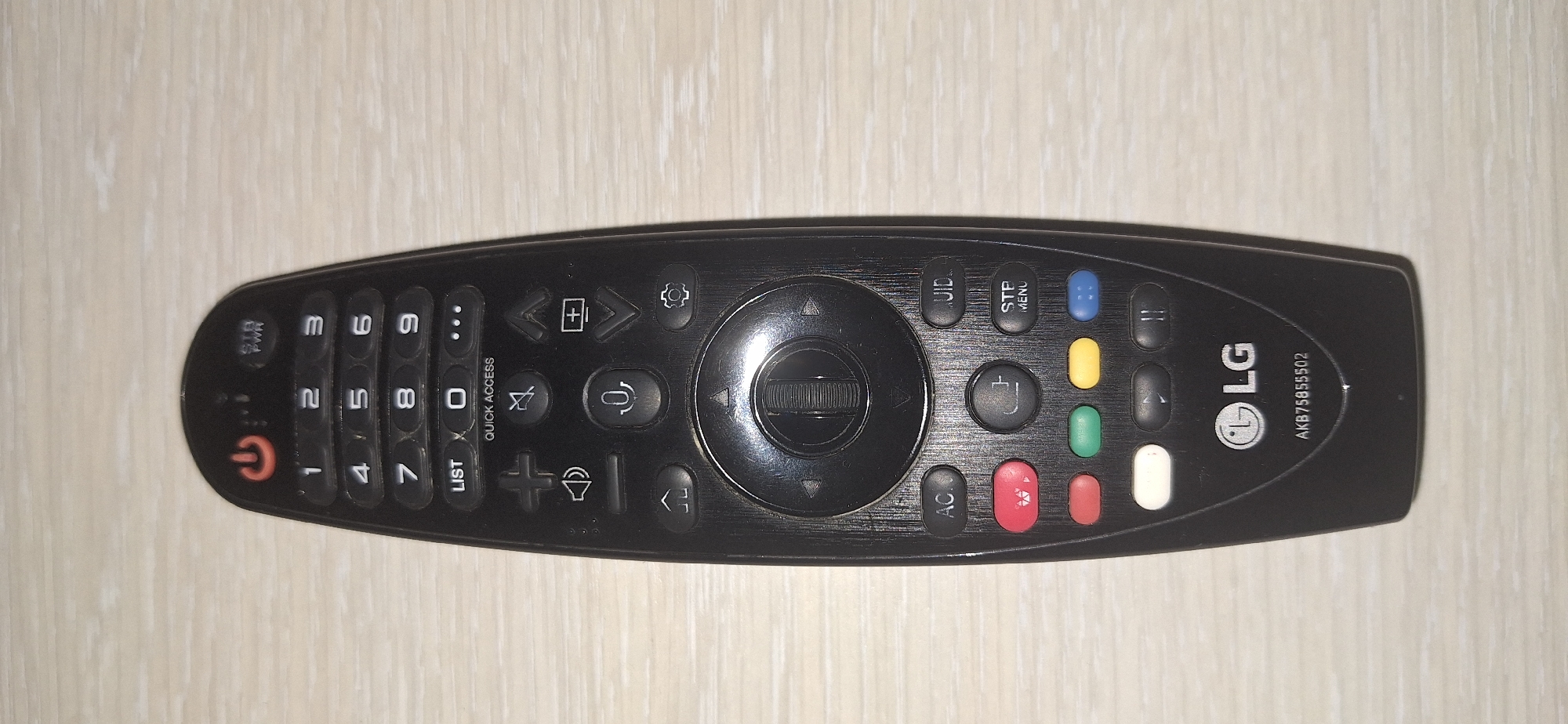 A black LG television remote control with various buttons is positioned on a light-colored wooden surface. The remote features a power button at the top, followed by a numeric keypad, and then a row of function buttons including "LIST", a square button, and three dots. Below this are buttons for volume control, microphone input, and navigation. At the bottom are colored buttons (red, green, yellow, blue) and playback controls, culminating in the LG logo and a model number "AKB75855502". The background is a close-up of a wooden tabletop. There are no people or any specific location markers that would indicate Bila Tserkva, Ukraine.