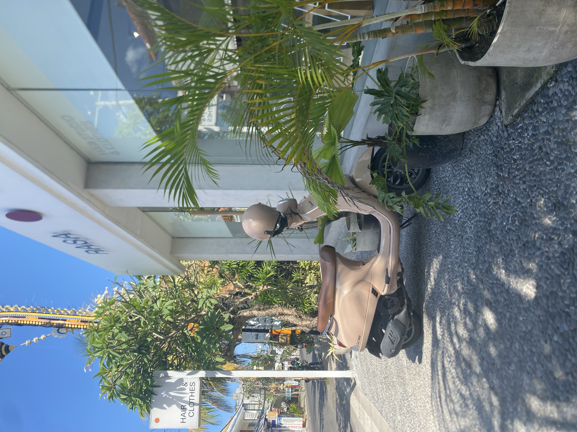 In the city of Mengwi, Indonesia, a light brown Vespa scooter is parked on a pebbled sidewalk in front of a building with large glass windows. The building has a sign that reads "RASA HAIR STUDIO & CLOTHES". Several potted plants, including palm trees and leafy green plants, are placed along the sidewalk in front of the building. A tall, ornate Balinese structure with yellow and orange accents is visible in the background, along with lush green trees and other buildings. The sky is clear and blue. The scooter has a helmet resting on its seat. A street is visible in the background with a few people and motorbikes. Sunlight casts shadows from the plants onto the sidewalk.
