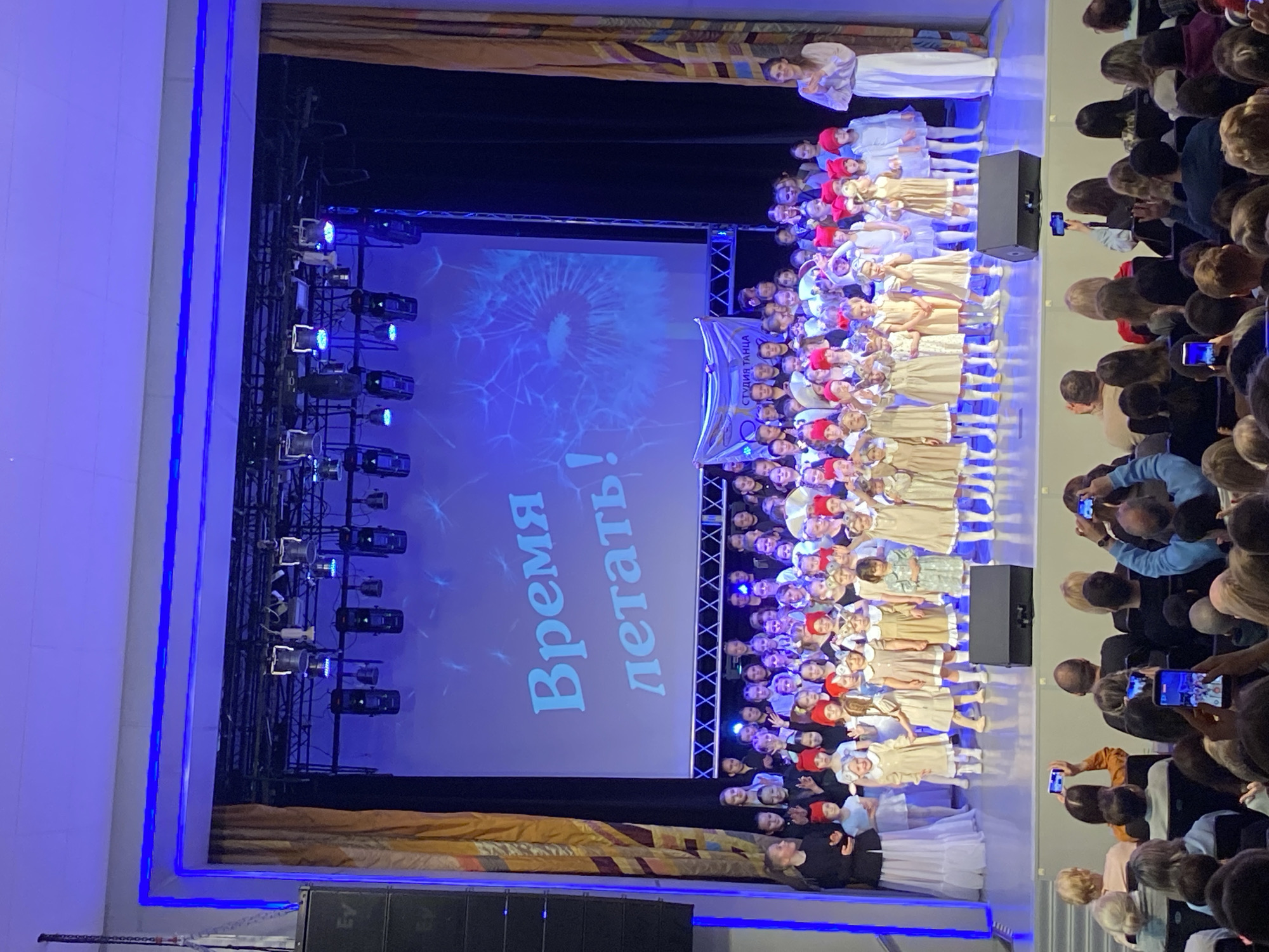Here is a description of the image:
The image depicts a stage performance, likely a children's dance recital, in what appears to be an auditorium setting. A large group of children, dressed in costumes (mostly light-colored dresses and some with red hats), are standing on the stage. Behind them is a screen displaying a dandelion-like image and the words "Время летать!" ("Time to Fly!"). The stage is equipped with lighting rigs visible on the left. An audience is visible on the right side of the image, many of whom are holding up phones to record or photograph the performance. Based on the banner held by the children with the studio name and location, the event takes place in Chaikovsky, Russia.