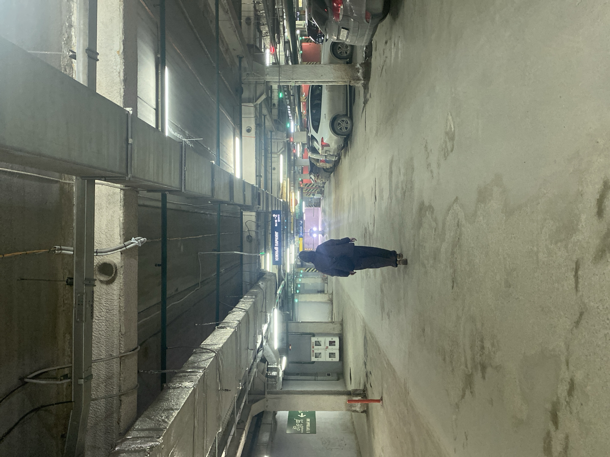 The image shows a view down a narrow parking garage lane, apparently in Yekaterinburg, Russia, based on signage. A single individual in dark clothing is walking away from the camera in the lane. Cars are parked on the left side. The right side is a concrete wall. Overhead is industrial ductwork. Lighting is from fluorescent or LED fixtures. The floor appears to be concrete. There are pipes and other utility infrastructure visible.