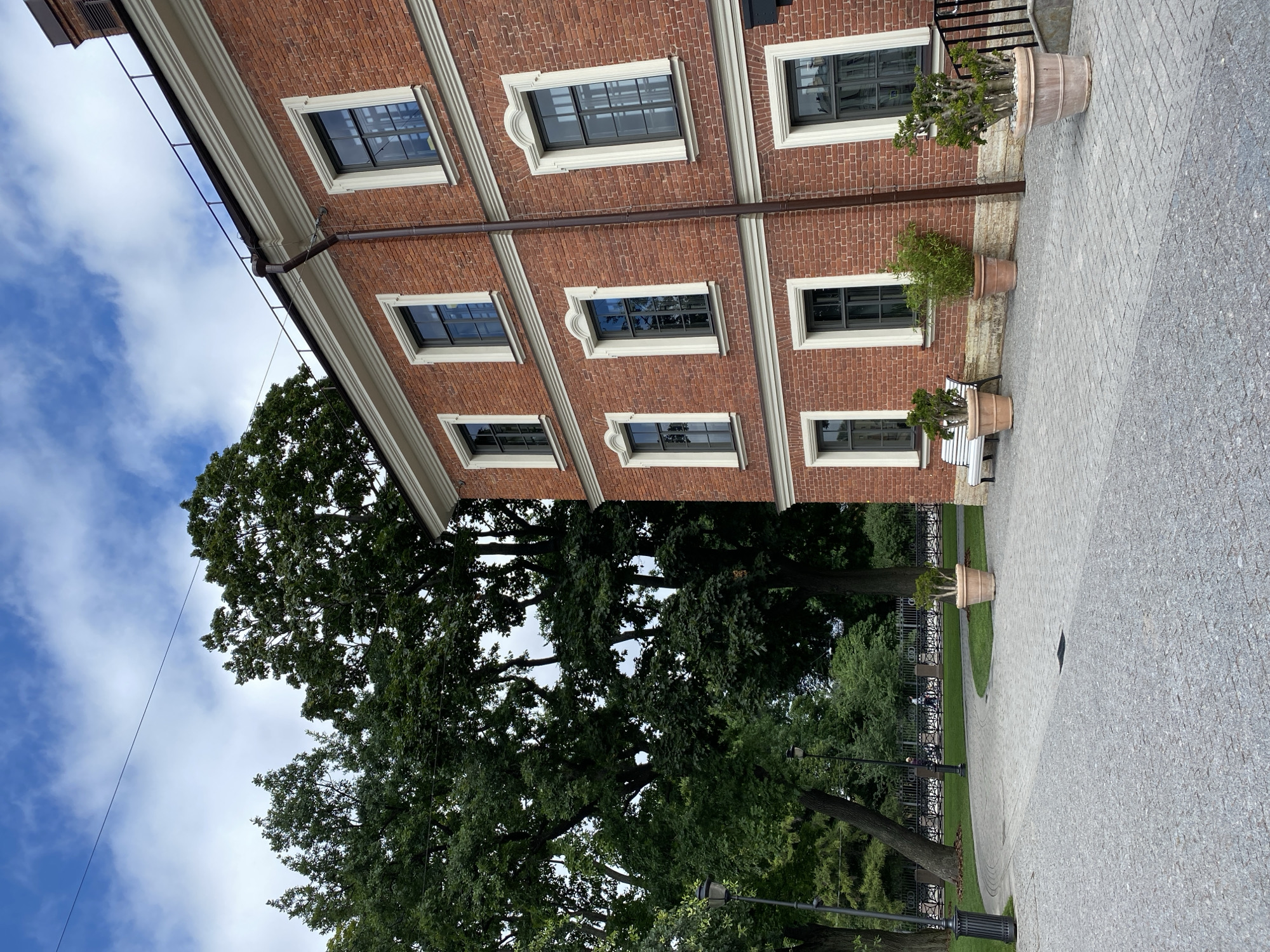 This is a photograph of the exterior of a red brick building, likely a historic structure, in Saint Petersburg, Russia. The building features multiple windows with dark frames and decorative white trim. In front of the building, a paved walkway is visible, made of small grey stones. To the left of the building are large, mature trees with lush green foliage, and a park area with a black metal fence. There are also several large potted plants placed along the walkway in front of the building. The sky is partly cloudy, with white clouds against a blue sky. The scene appears to be a public space, possibly a park or a courtyard adjacent to a cultural institution or administrative building, given the architectural style and the presence of trees and park features.